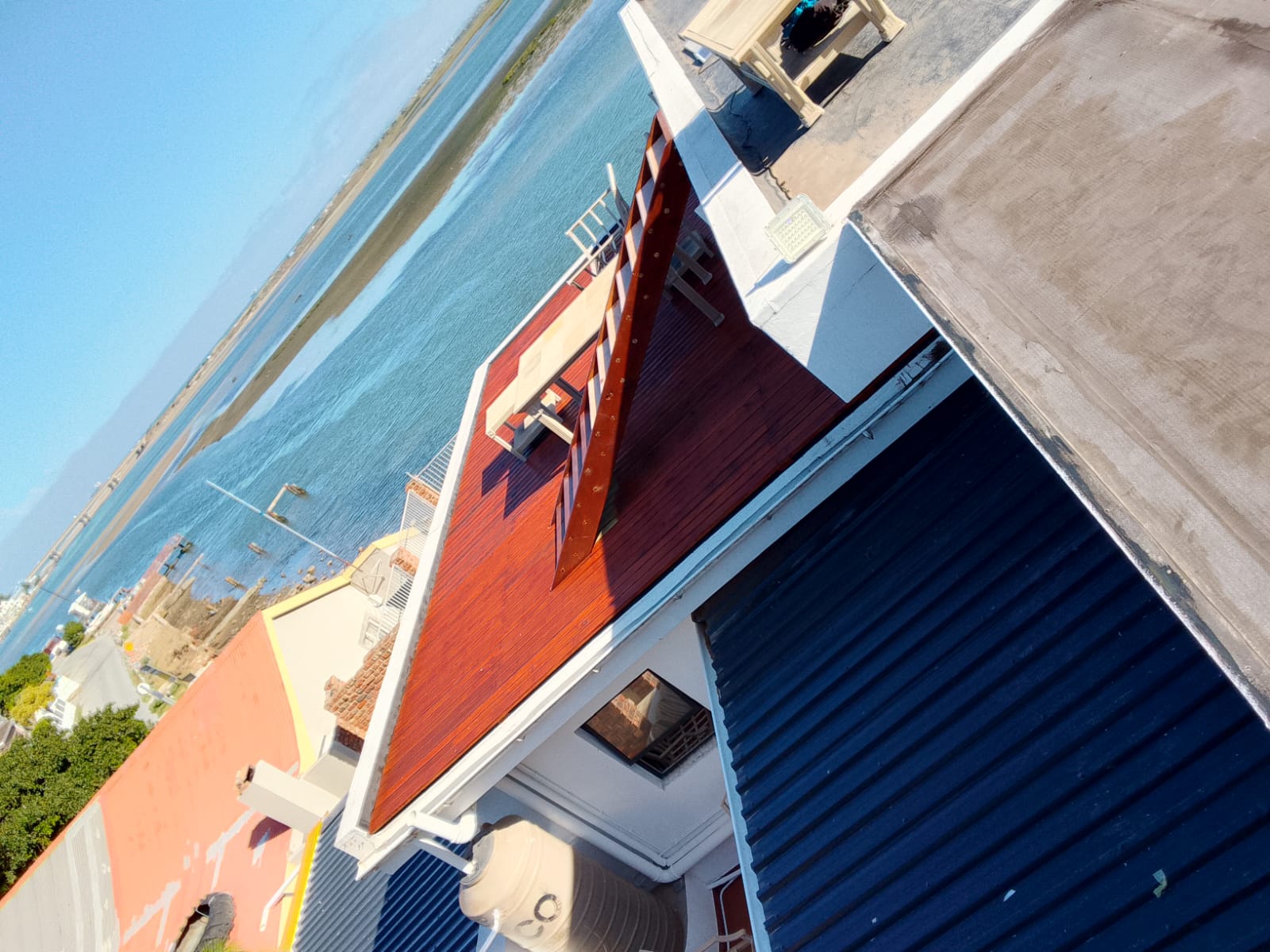 Dramatic tilted aerial of the complete mahogany deck with open water and estuary behind, white parapet walls and PVC gutter, water tank and corrugated iron roof below