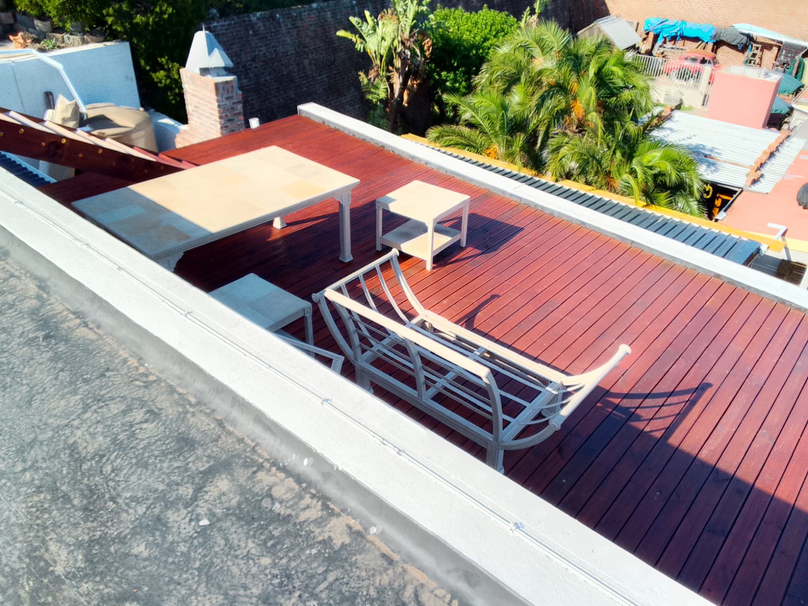 Overhead aerial of finished mahogany rooftop deck with white lounge chair and two side tables, lush palm trees and greenery behind