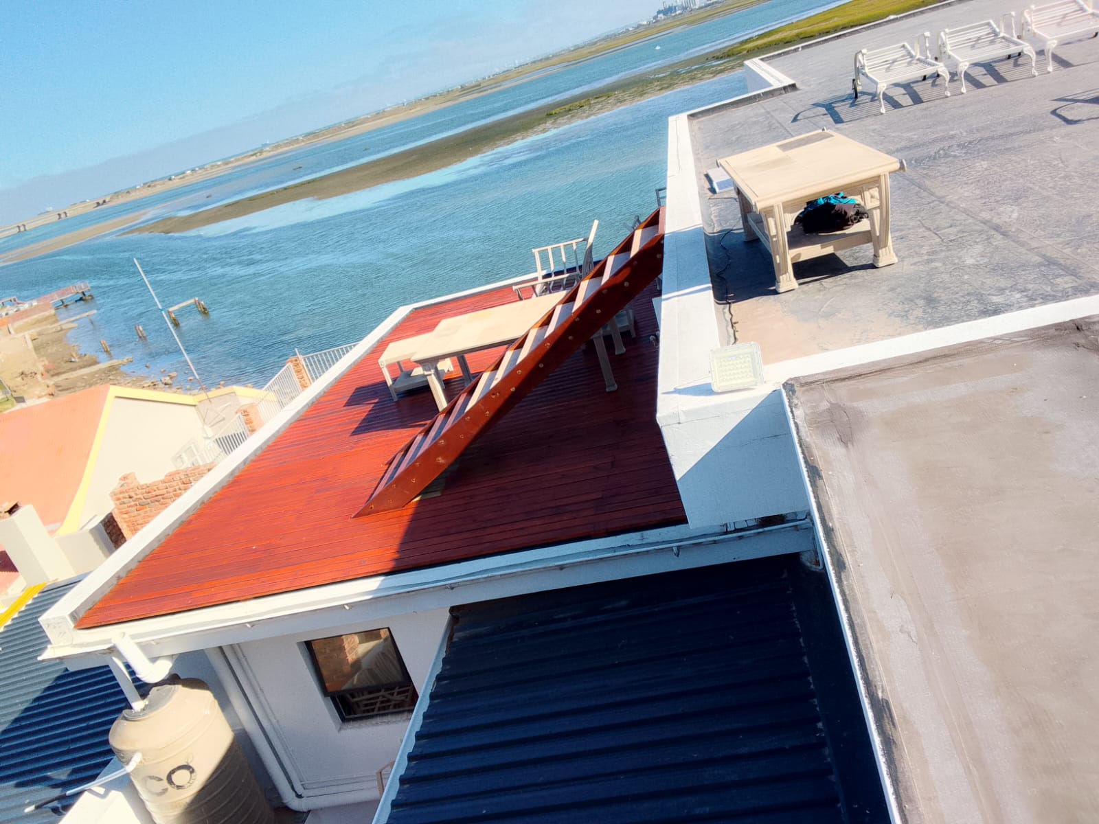 Elevated view of deck mid-staining showing red and natural pine sections side by side, lagoon and estuary visible in background, chairs placed on stained portion