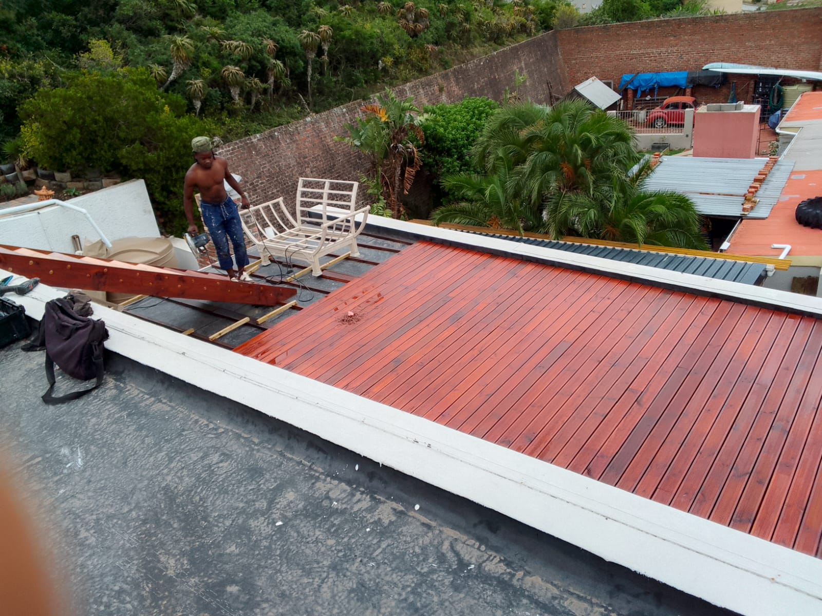 Worker using power sander on deck surface during staining, aerial view showing majority stained red, chairs moved to side, green hillside background