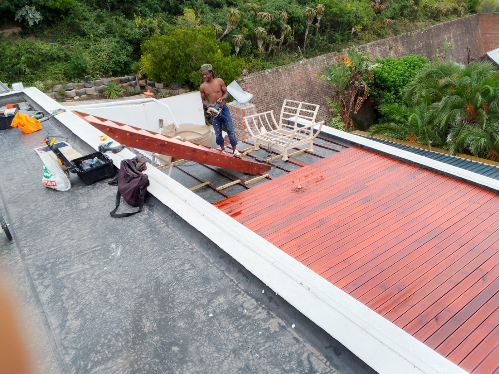 Worker smiling on deck during staining process, red and natural timber sections visible, green hillside and lush plants behind
