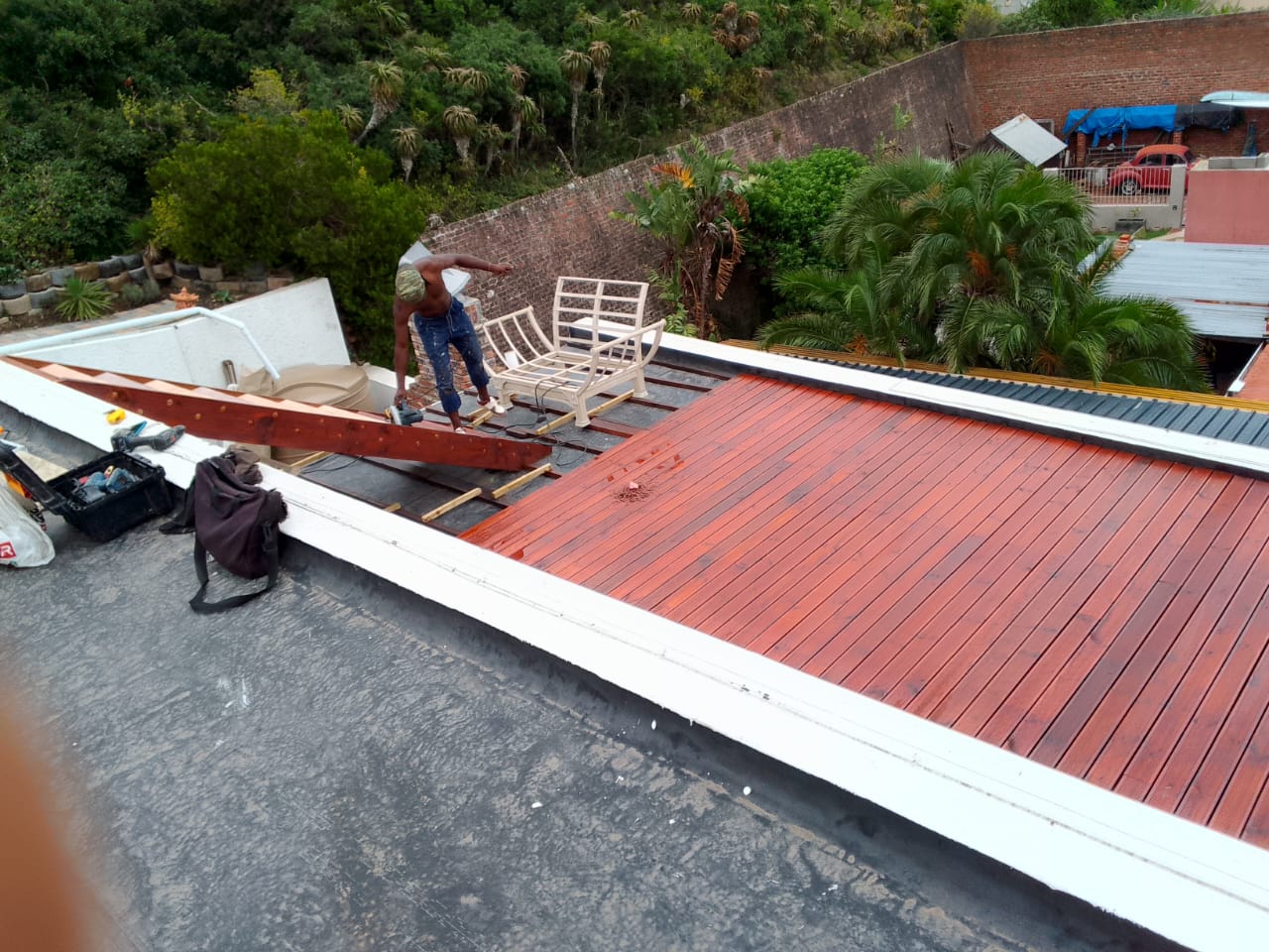 Worker standing on stained deck with power tool, rear half still natural pine, bird's-eye view with lush green hillside and aloes behind