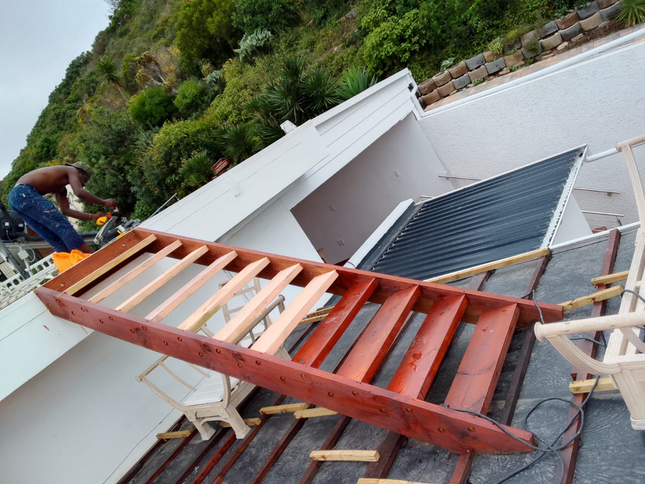 Close-up overhead of treated red timber deck subframe — main beams and raw pine cross-joists bolted together, worker cutting at left, solar panels and hillside visible