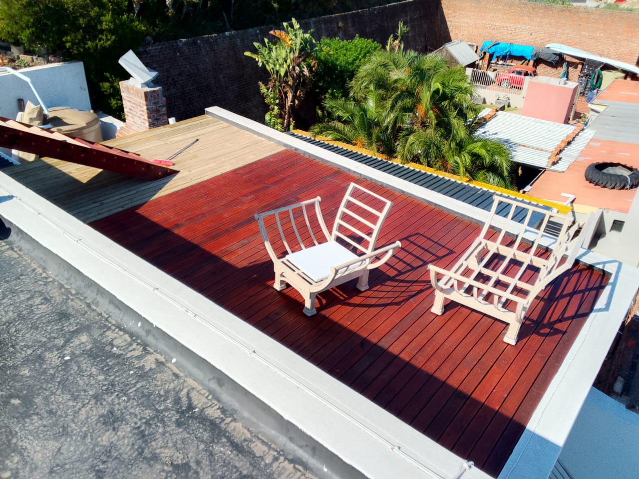 Aerial view of near-complete rooftop deck with two white chairs placed on rich red stained boards, natural timber section at back corner still remaining, palm trees visible