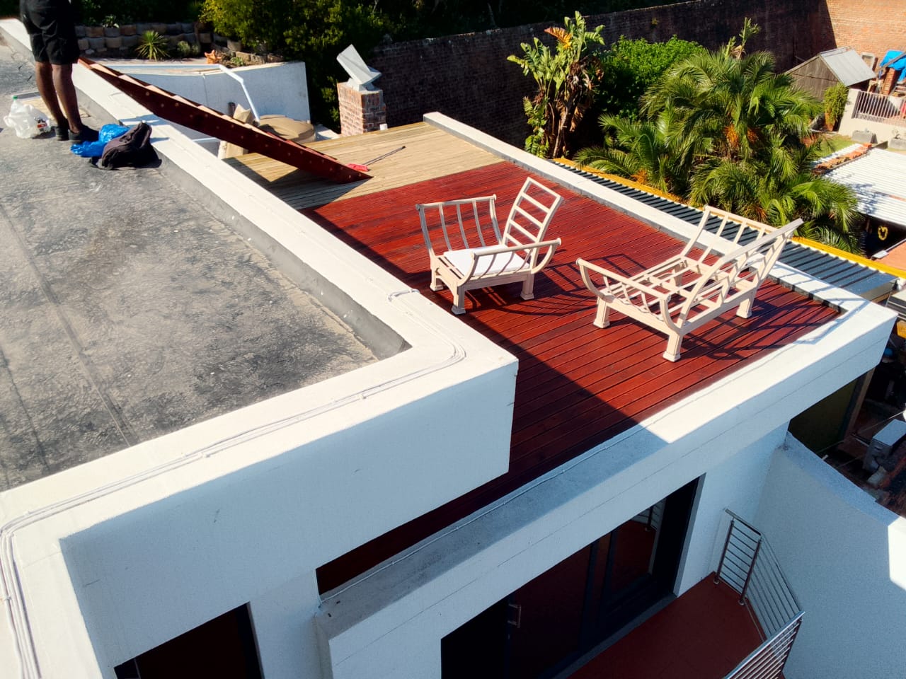 Aerial view of mahogany-stained deck with two white recliner chairs, unfinished natural timber section visible at far corner, garden behind