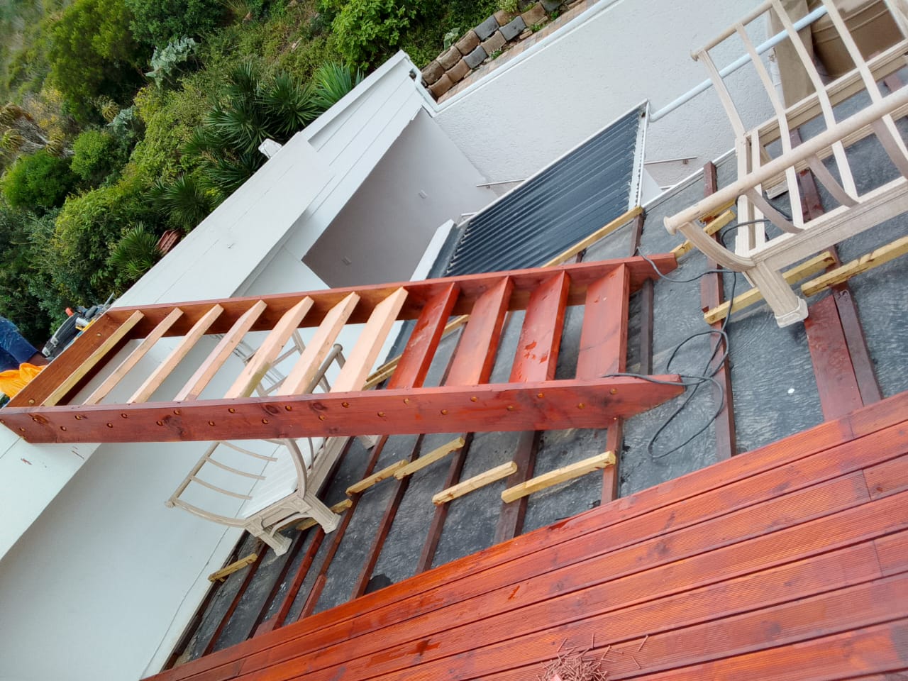 Close-up of stained red deck beam and cross-members being assembled, partial decking boards started at right, solar panel and chairs visible