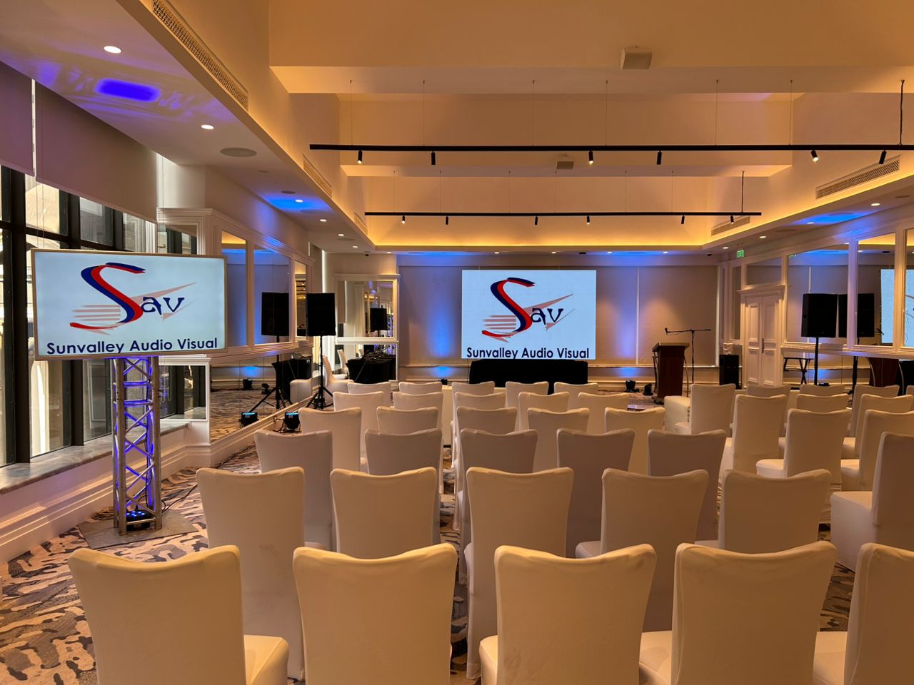 Professional event venue with dual LCD screens displaying Sunvalley Audio Visual branding, warm golden lighting with blue accents