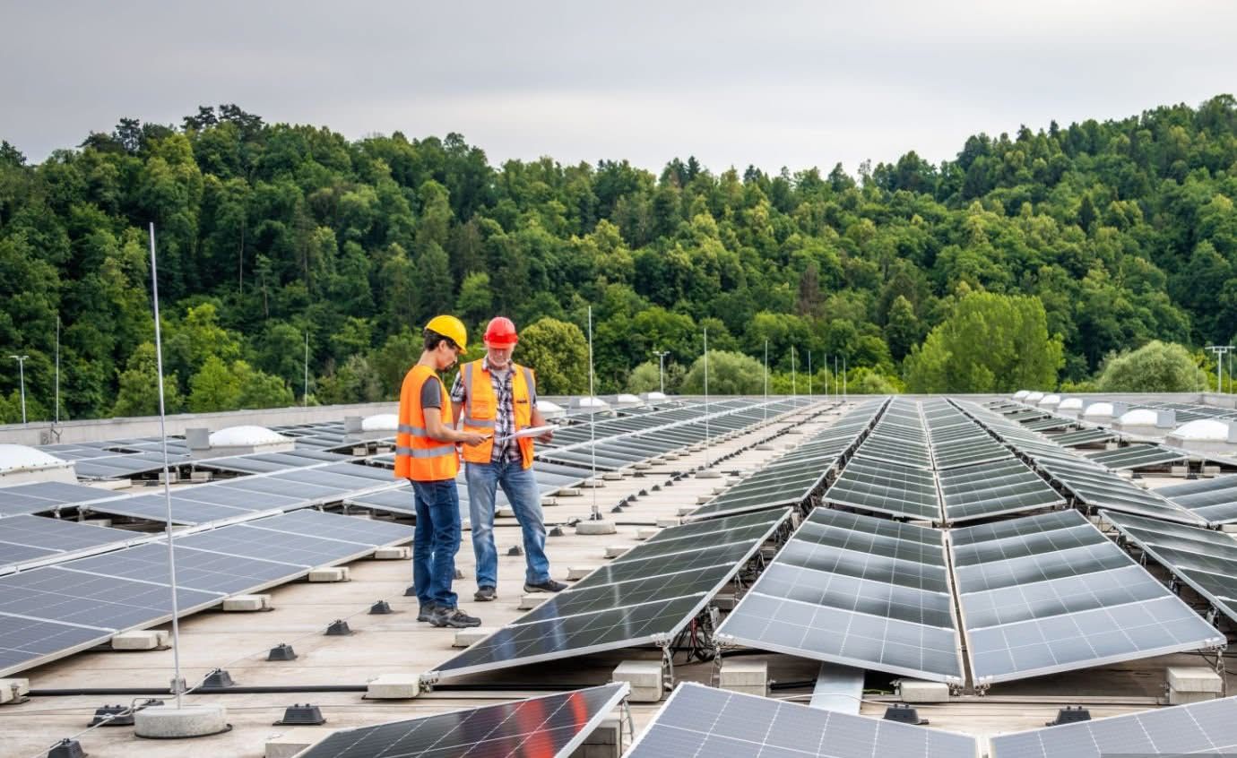 Due tecnici con giubbotti arancioni e caschi ispezionano impianto fotovoltaico su tetto industriale