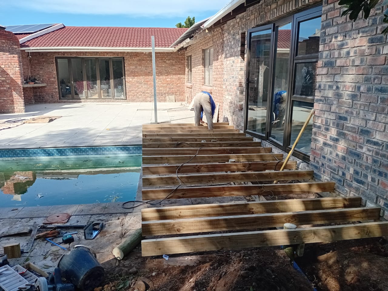 Worker building deck subframe with timber joists near pool, brick house with solar panels visible