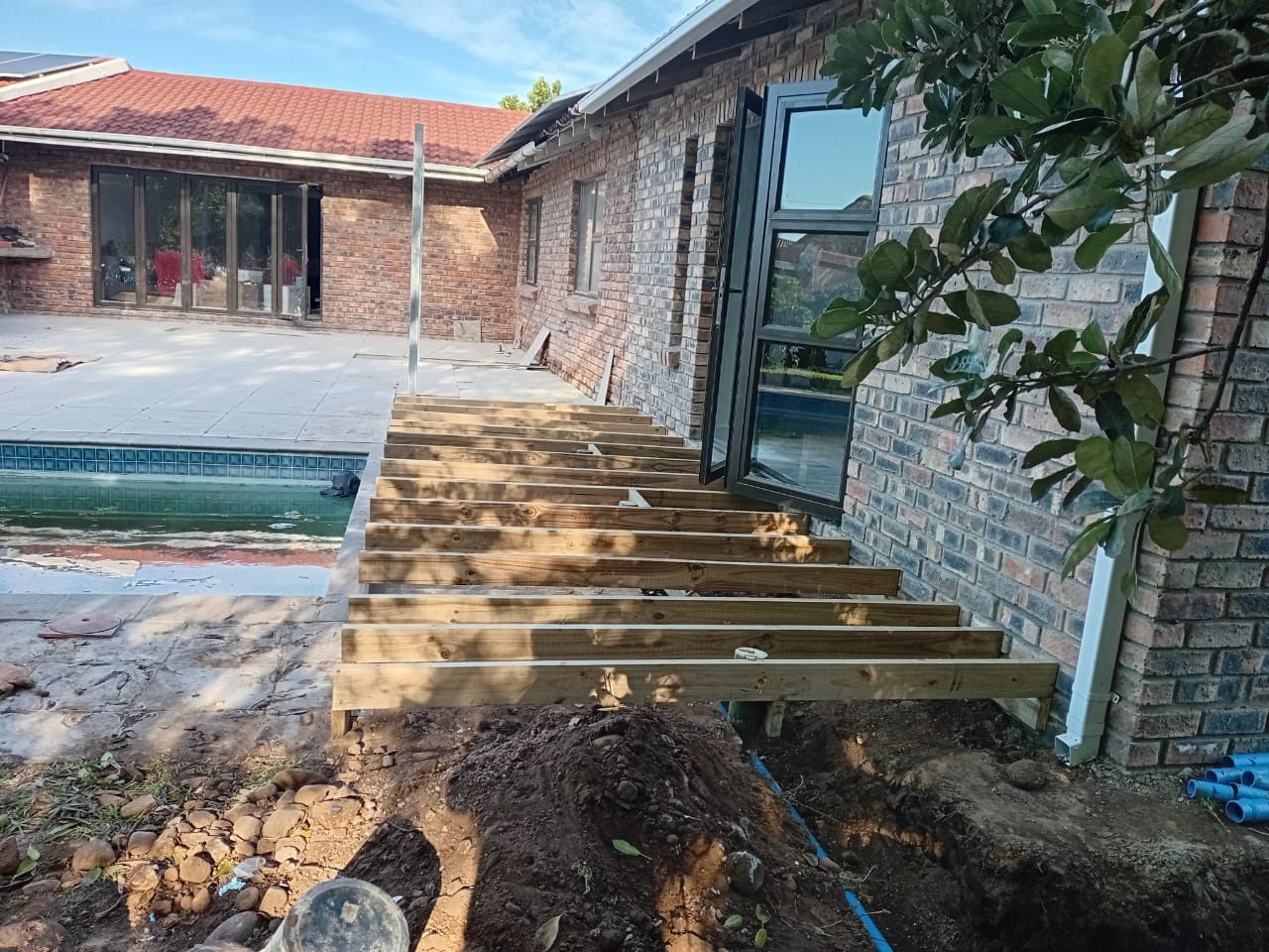 Wooden deck joists being installed next to pool, timber frame extending from house, excavation visible