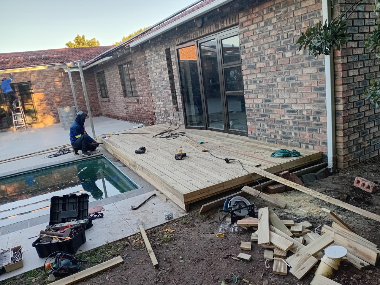 Worker installing wooden deck planks next to pool, tools and materials scattered, brick house in background