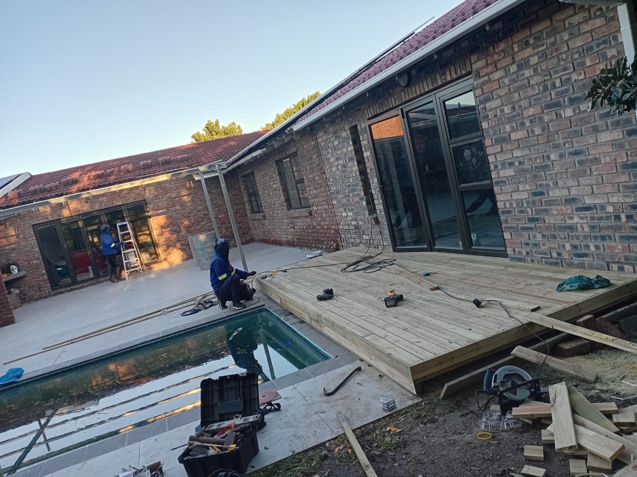 Wide view of deck installation with worker installing boards, pool and brick house visible, tools and sawdust around