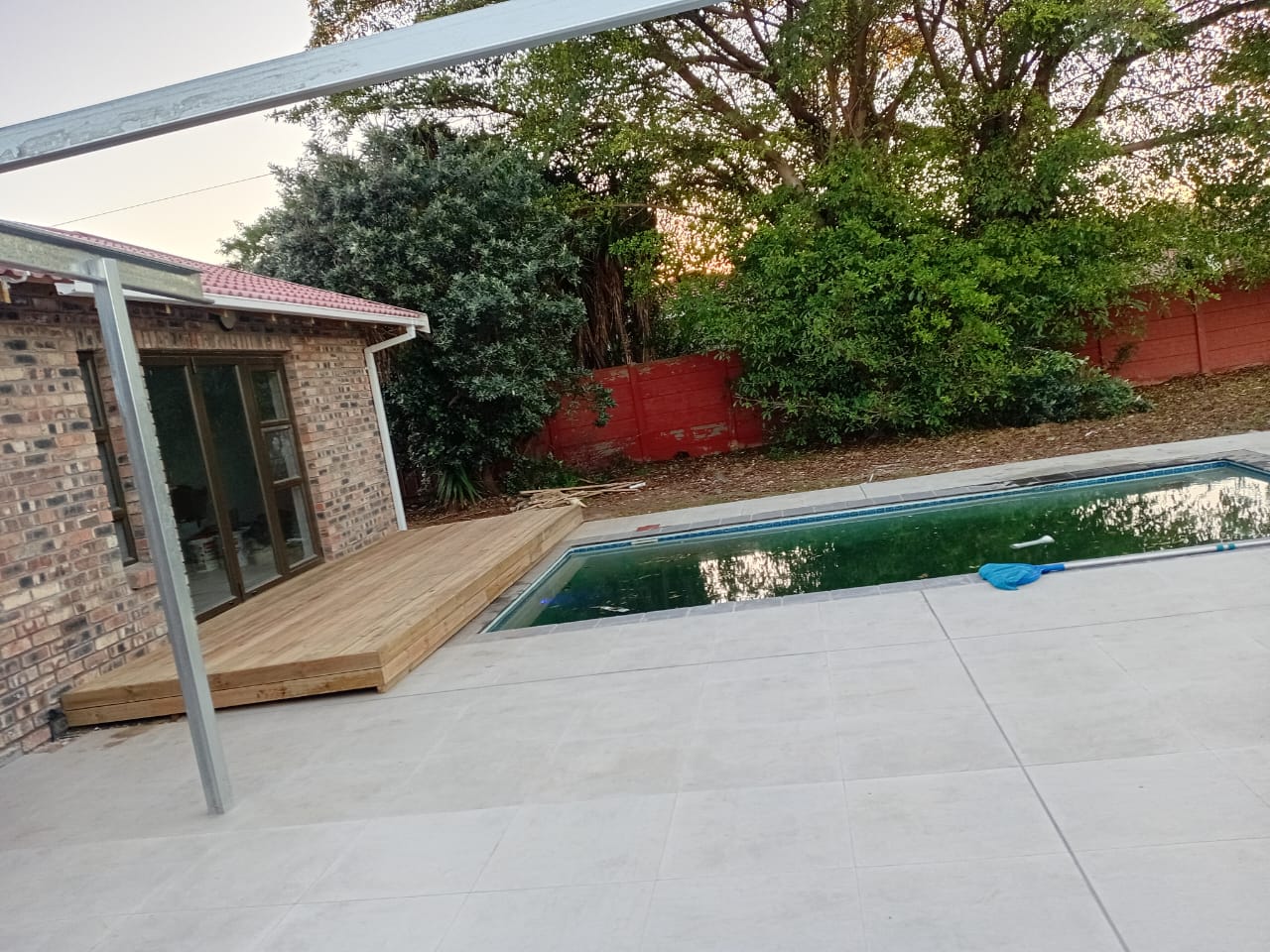 Completed wooden deck next to swimming pool with pergola frame overhead, brick house and garden visible
