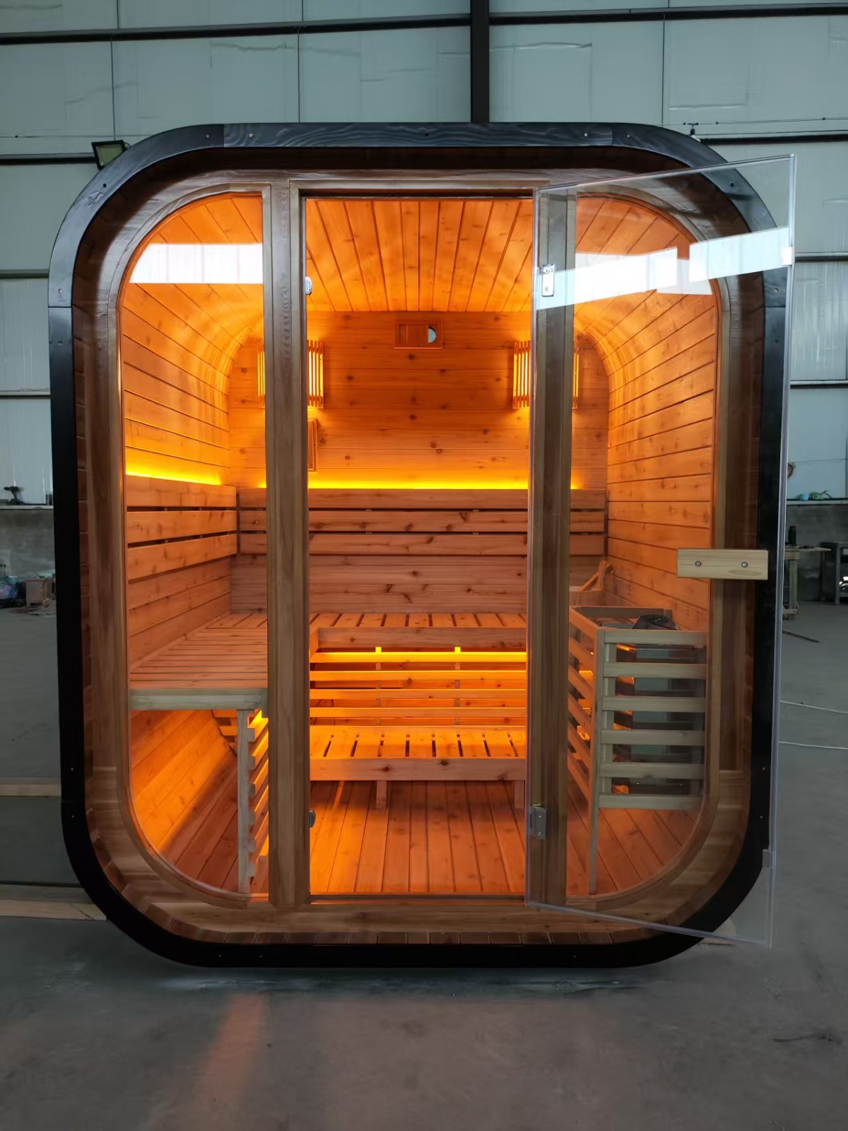 Front elevation of the finished cedar sauna with the glass door open, showing bench seating and warm lighting