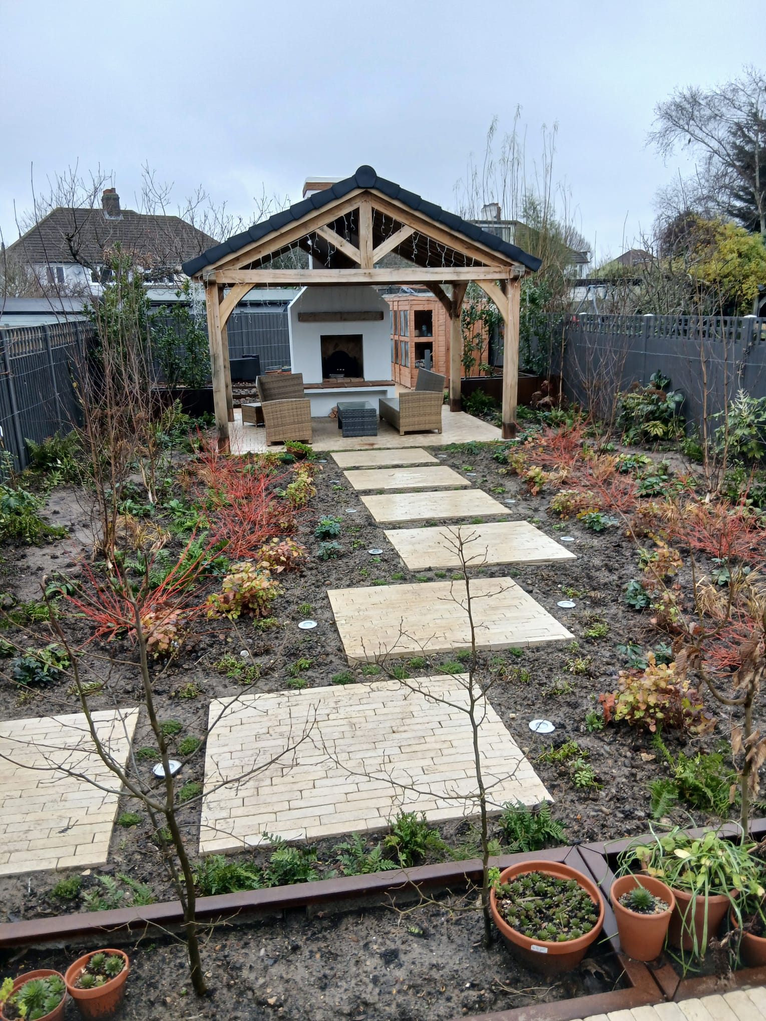 Oak gazebo with a white outdoor fireplace and timber store, fronted by a stepping-stone stone path through freshly planted beds