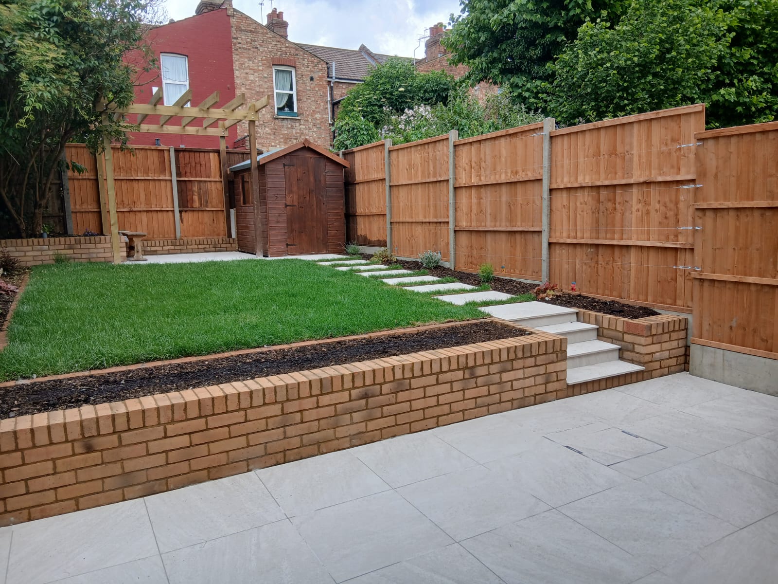 Tiered garden with a brick retaining wall and steps, a level lawn, porcelain-paved patio, timber shed and new fence panels