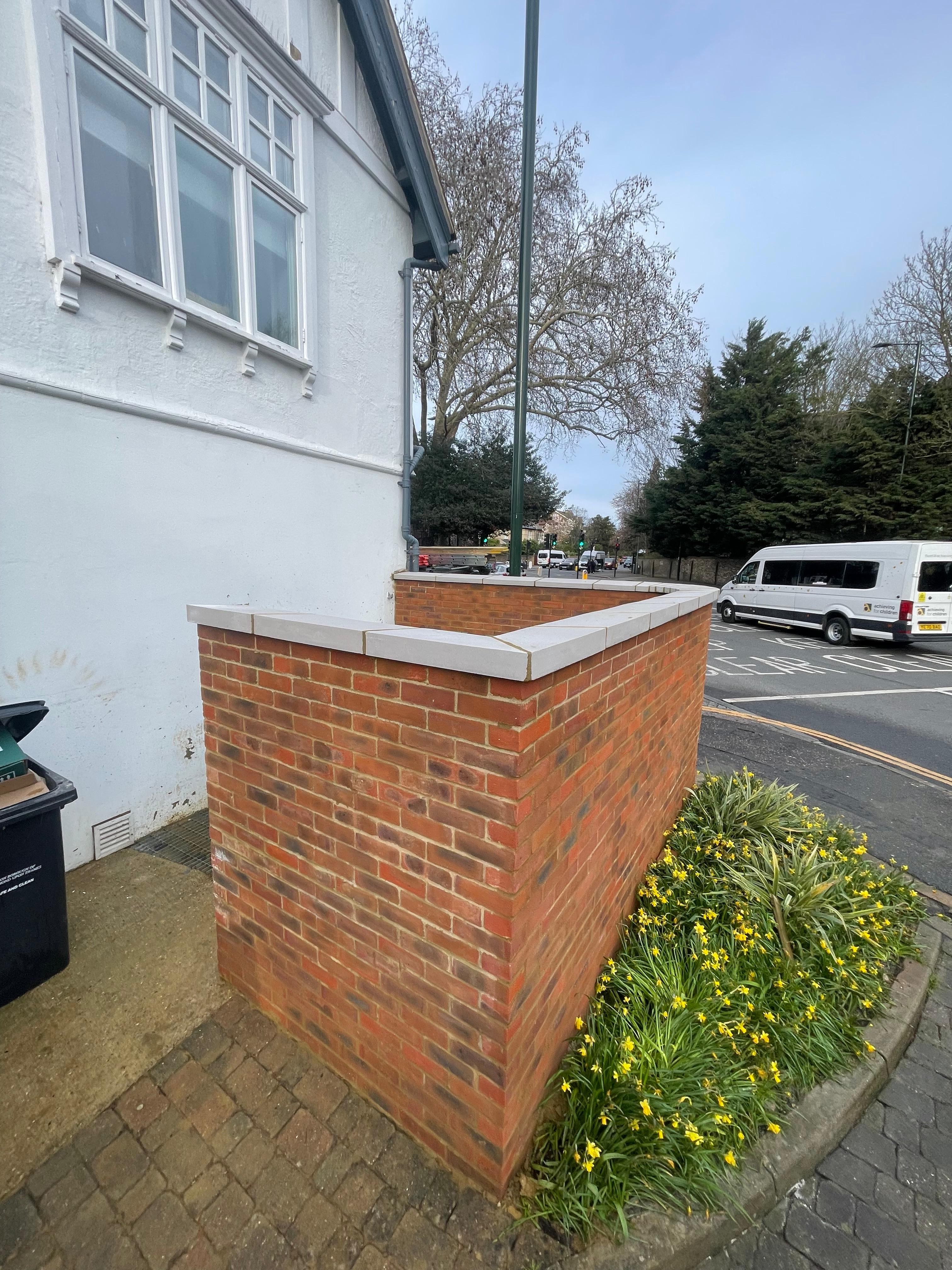 Red-brick corner boundary wall with crisp white stone coping beside a rendered period house and a bed of daffodils