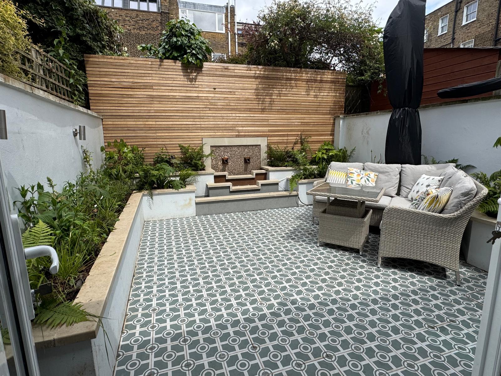 Contemporary courtyard with patterned encaustic floor tiles, a tiled water feature, cedar slatted screen, raised planting beds and a rattan sofa