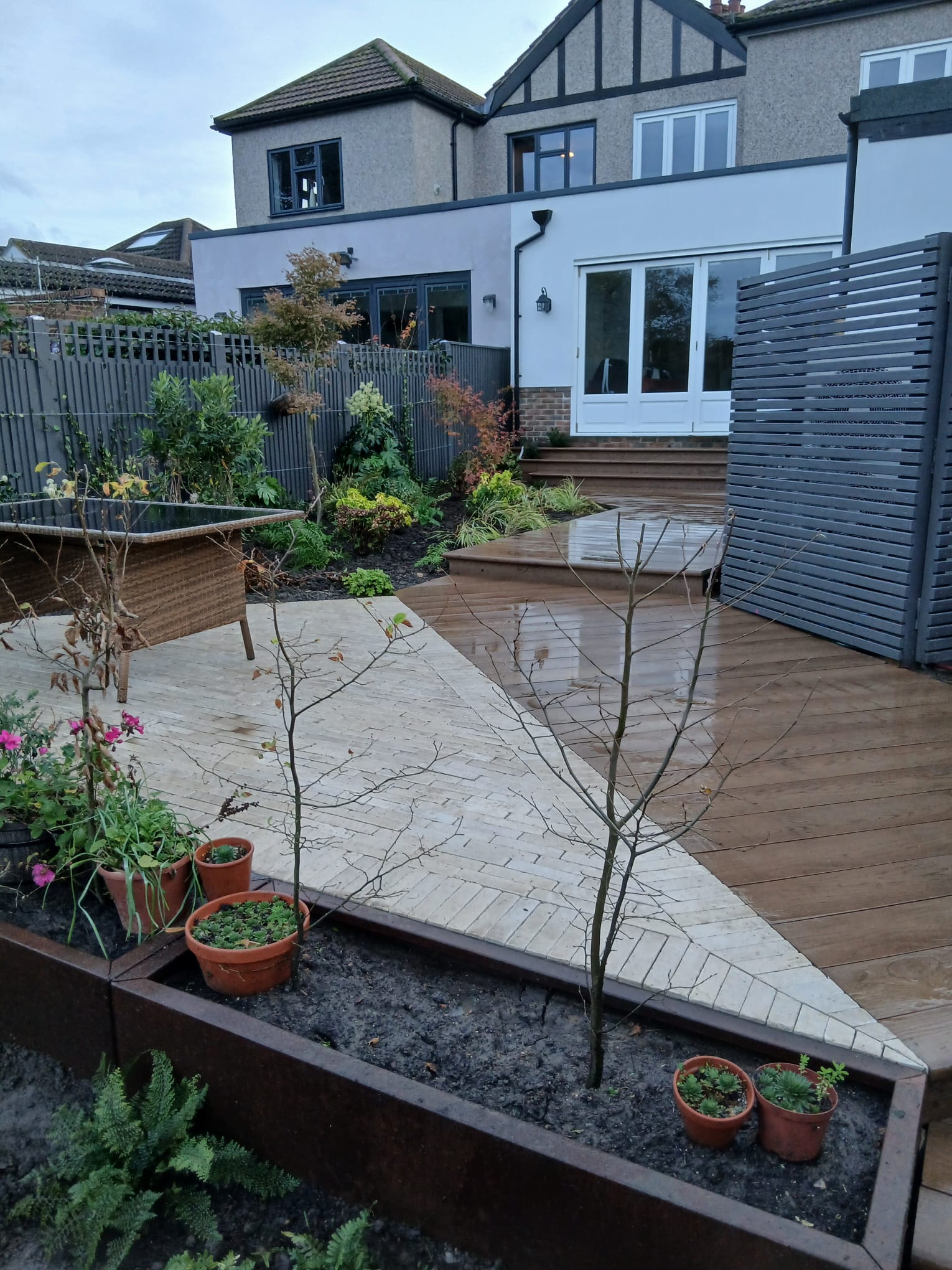 Rear garden with composite decking, a raised dining terrace, grey slatted privacy screen and planted borders leading to a rendered house extension
