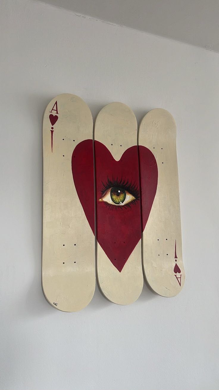 Ace of Hearts playing card with a painted green eye inside a large red heart, spread across three vertical skateboard decks mounted on a wall