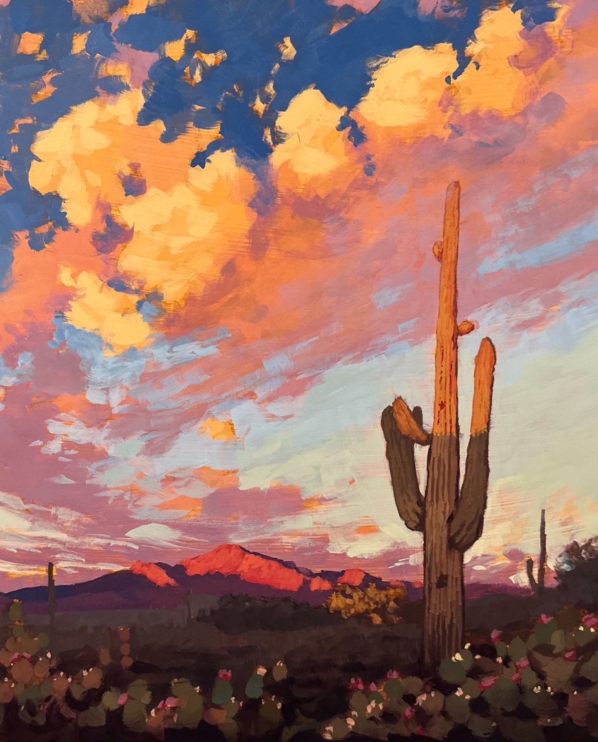 Tall saguaro cactus against a dramatic orange-gold-purple sunset sky with red mountain peaks and wildflowers