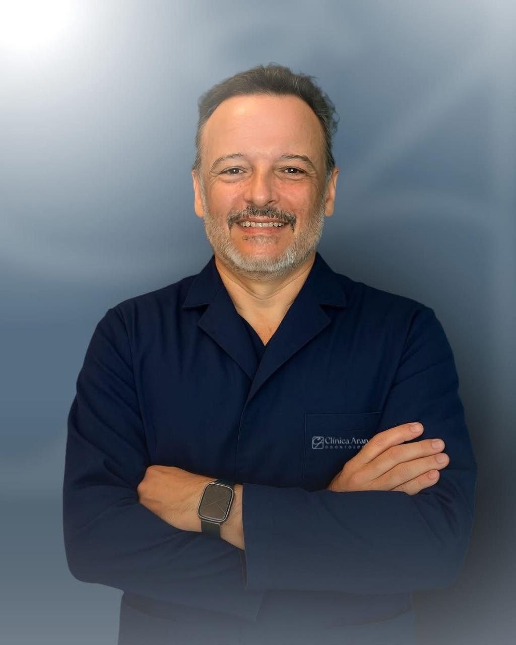Dr. André Arana com os braços cruzados e sorrindo, jaleco azul da Clínica Arana