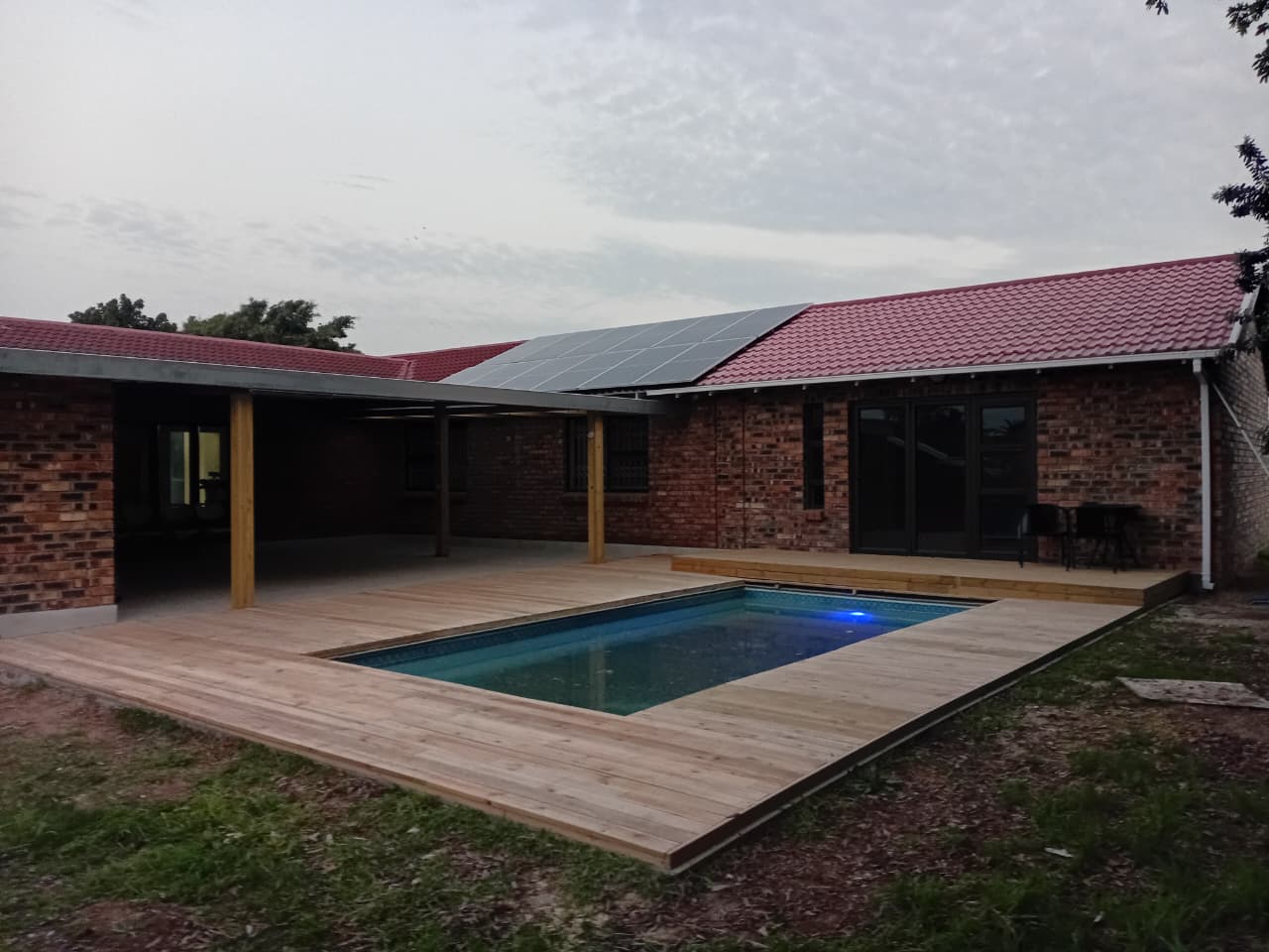 Poolside timber deck Gqeberha — completed natural pine deck surrounding rectangular pool, blue pool light glowing, brick house with timber pergola posts
