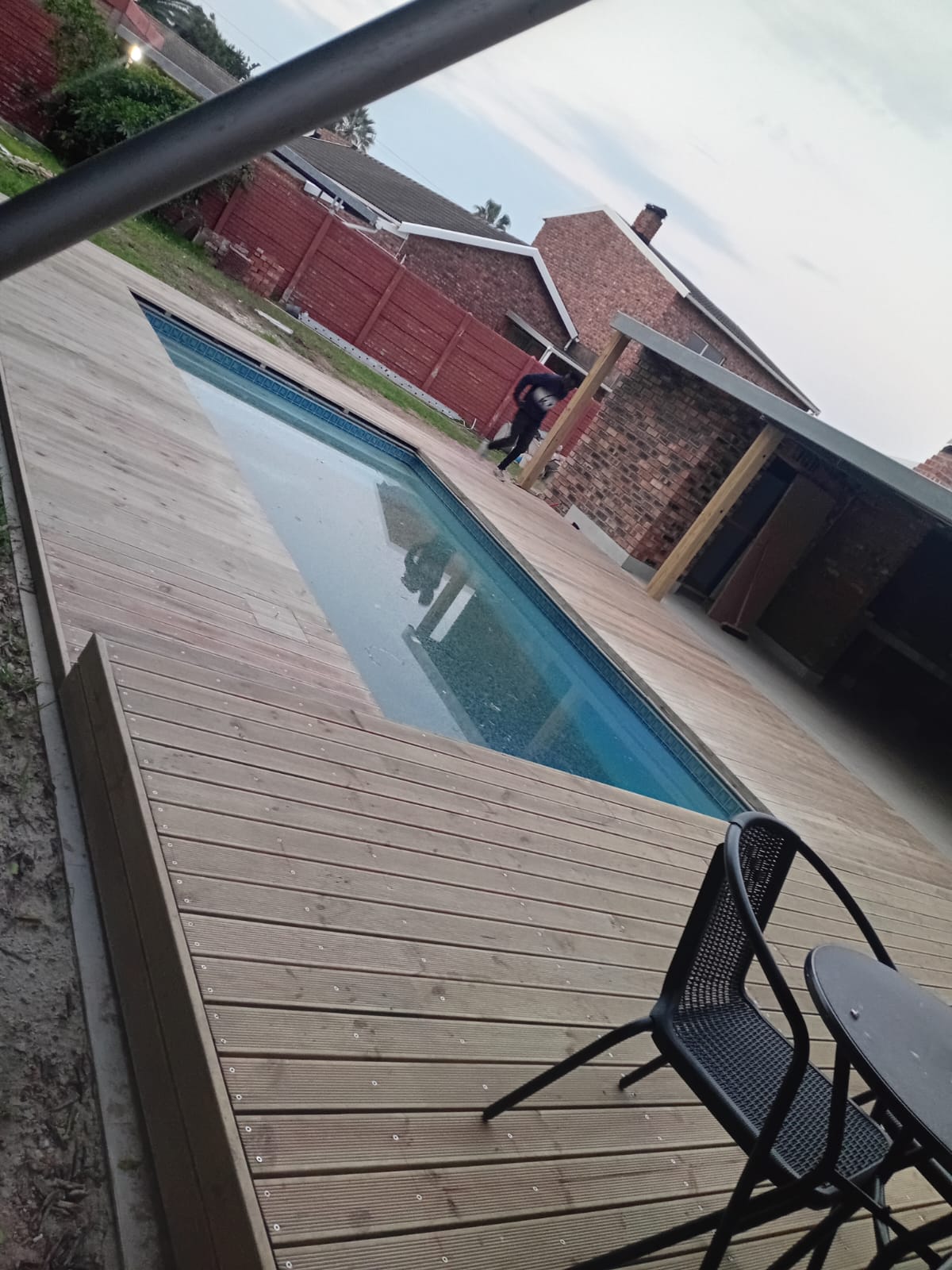 Close-up of finished pool deck boards with screw fixings, outdoor furniture placed on deck, pool and brick boundary wall visible