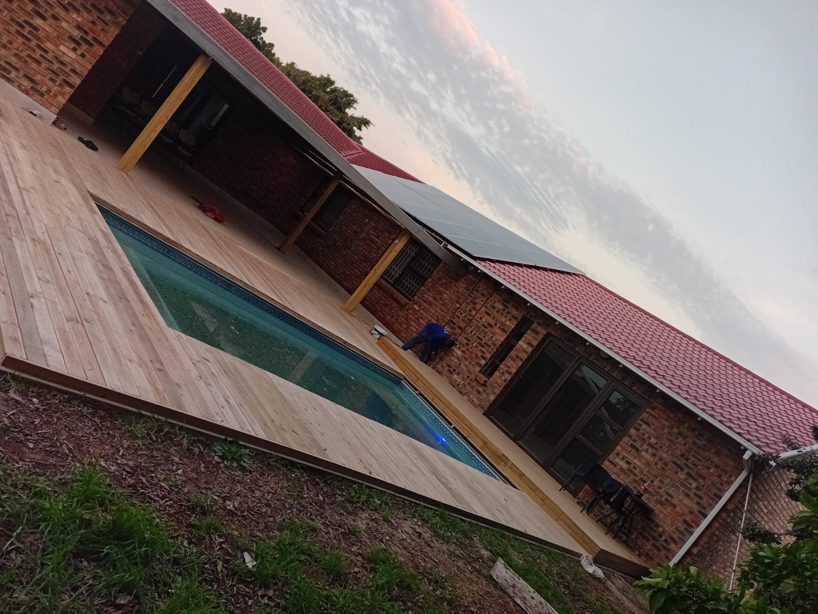 Elevated view of timber deck surrounding pool — full deck footprint visible, worker finishing boards, red tile roof and solar panels on brick house