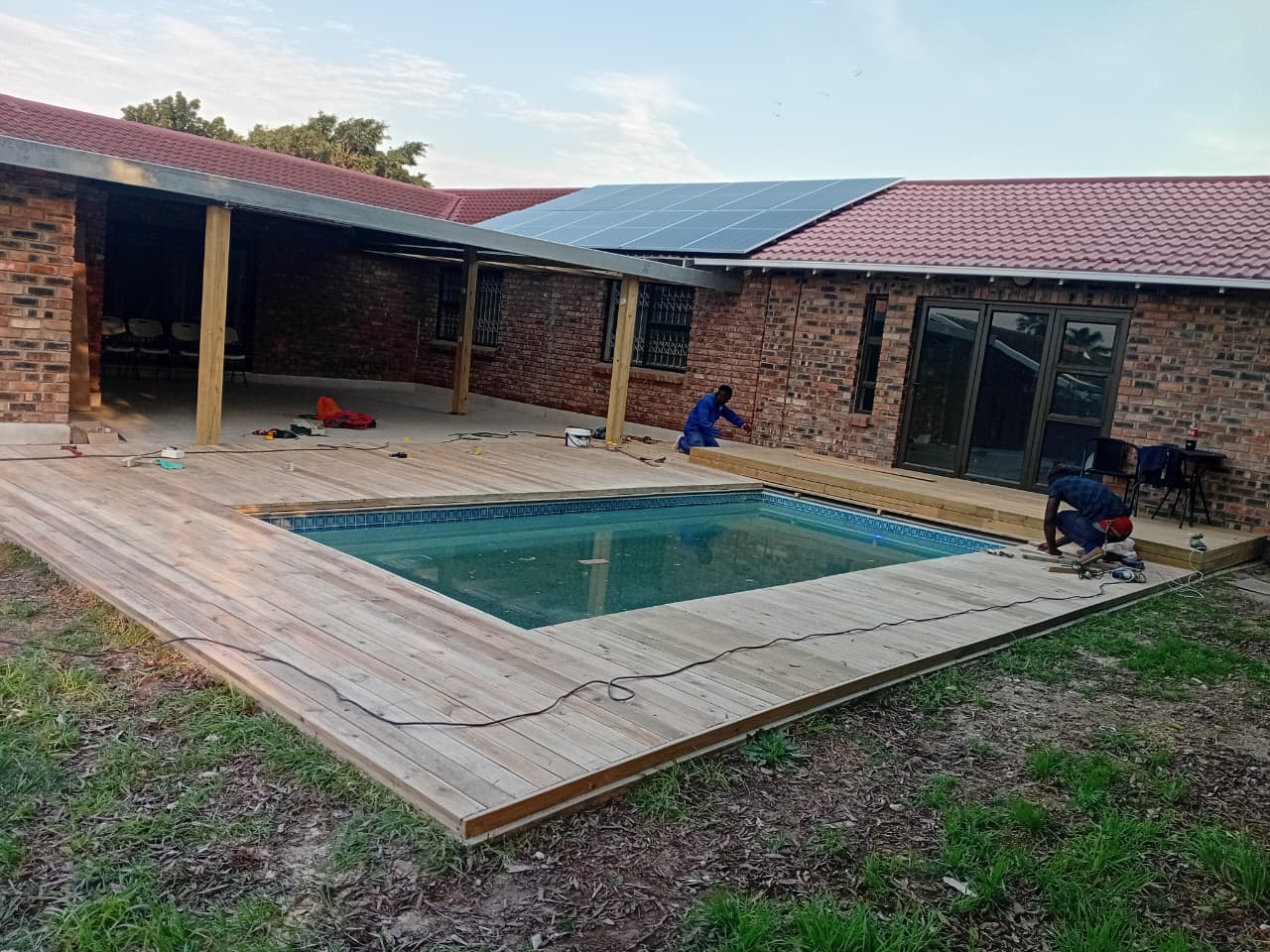 Wooden deck installation Port Elizabeth — two workers laying final pine boards around swimming pool, pergola structure and solar panels visible
