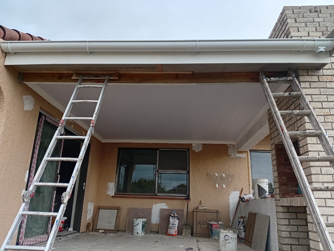 Flat roof soffit installation Port Elizabeth — white plasterboard ceiling being installed, metal ladder, brick and window frame visible