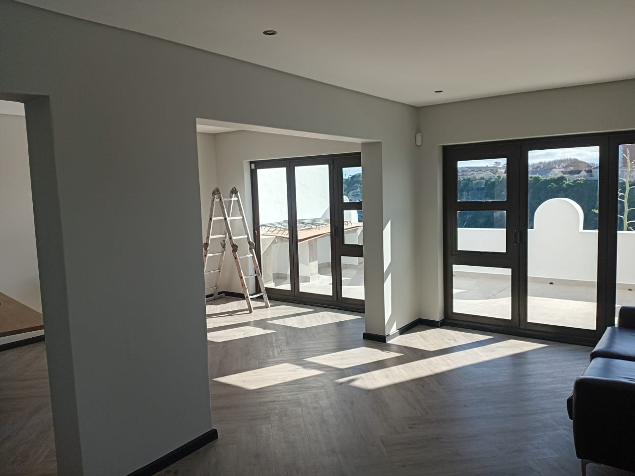 Interior painting Gqeberha — bright open plan lounge with light grey walls, two sets of dark-framed doors to terrace, herringbone floor, city skyline view