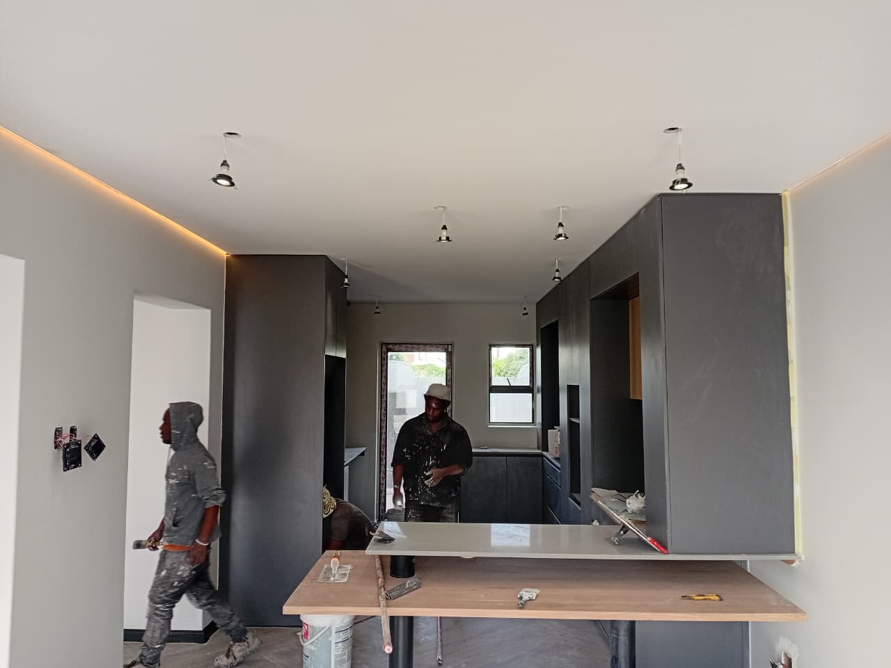 Interior painting Gqeberha — Osflex Construction painters working in kitchen with white ceiling, amber LED strip cove, charcoal cabinetry and pendant lights