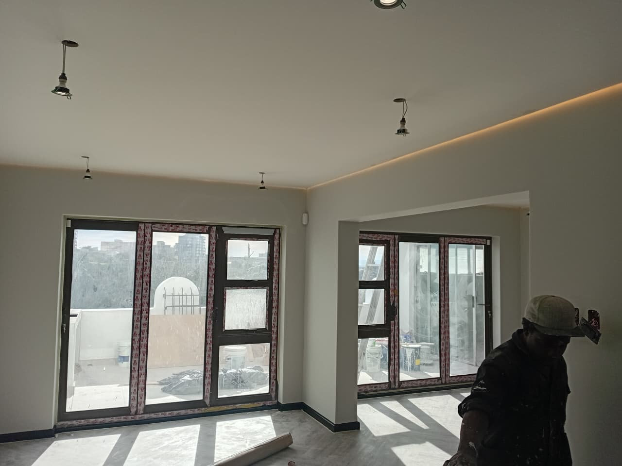 Interior painting Gqeberha — room with freshly painted cream walls, amber LED cove just visible, two large dark-framed windows, painter finishing window reveals