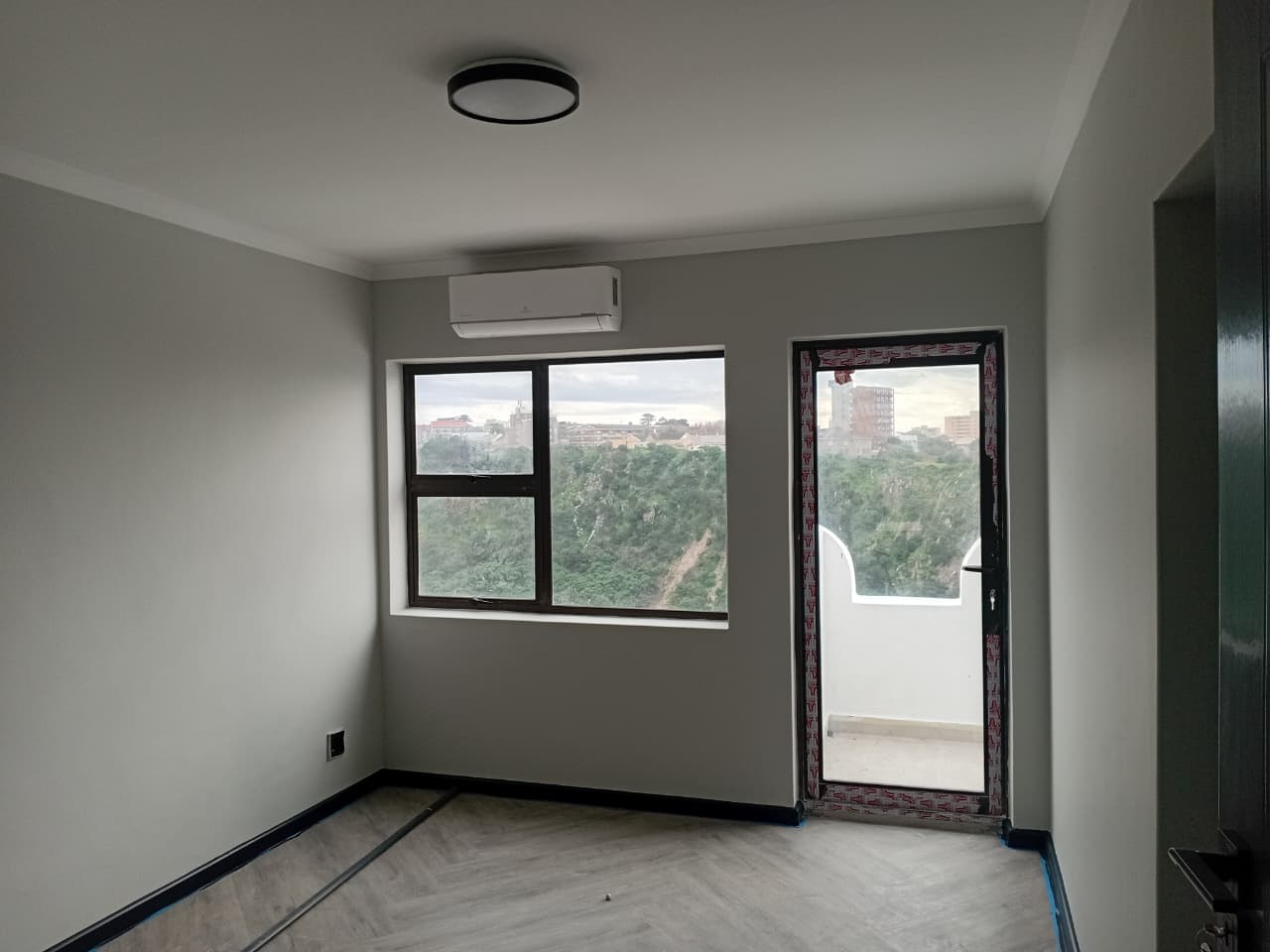 Interior painting Gqeberha — bedroom with grey painted walls, white cornice, round black ceiling light, air conditioning unit, balcony door with treetop view