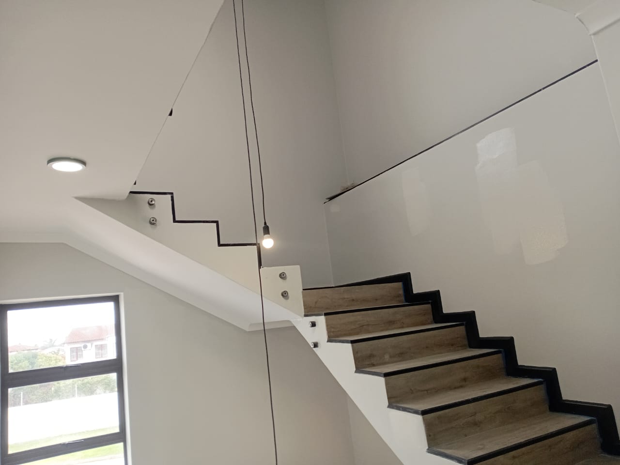 Interior painting Port Elizabeth — close-up of floating staircase with glass balustrade, white painted stringer wall, pendant bulb light, garden view window