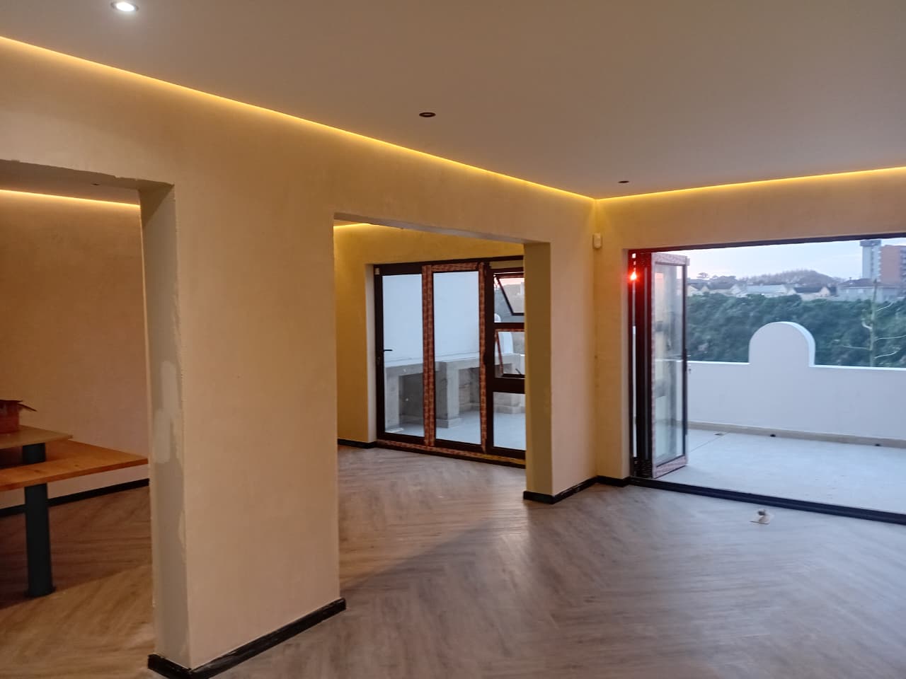 Interior painting Port Elizabeth — open plan lounge with warm amber LED cove lighting, cream walls, two sets of folding doors opening to balcony with treetop view