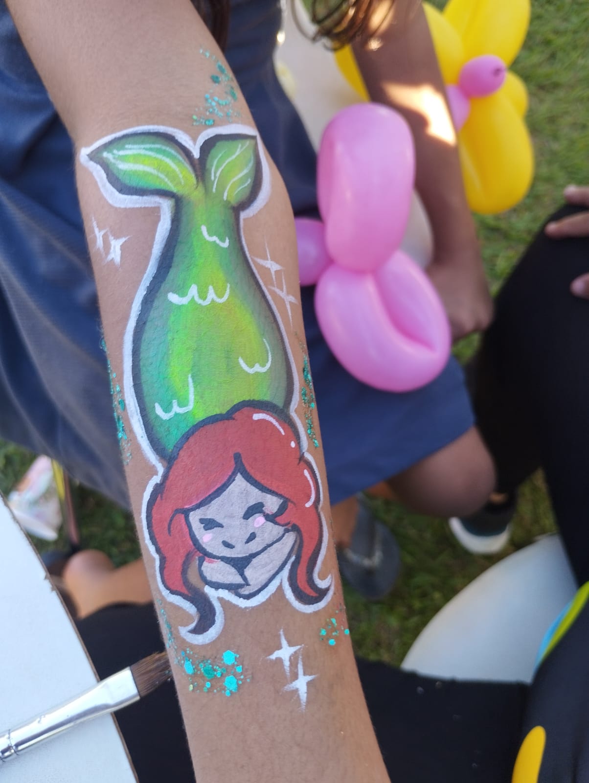 Pintura facial de sereia no braço de uma criança ao lado de balões modelados