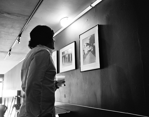Person standing in dark interior gallery space viewing framed black and white photographs on wall with track lighting