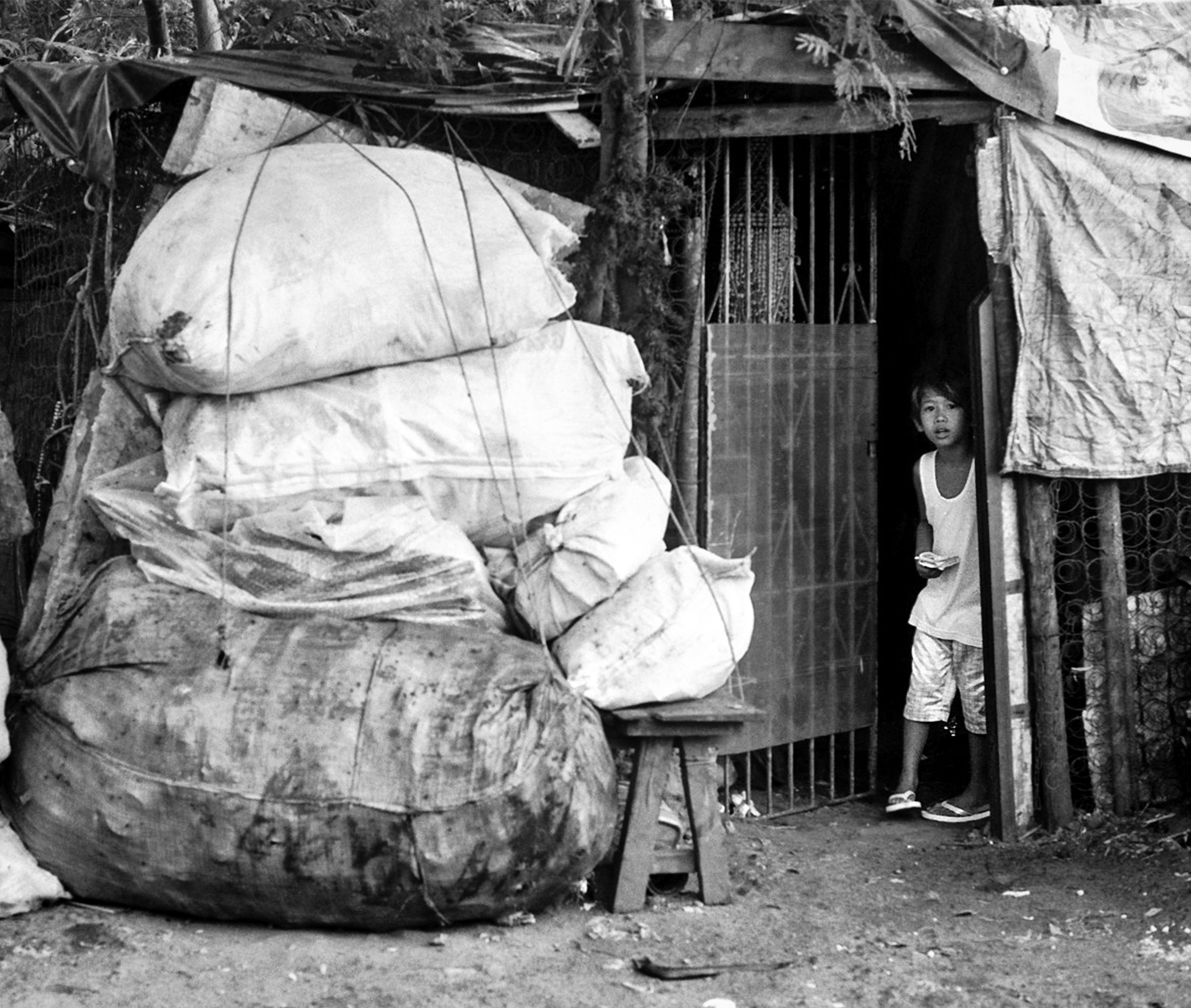 Young woman standing in doorway of modest dwelling with large stacked white sacks and hanging fabrics outside