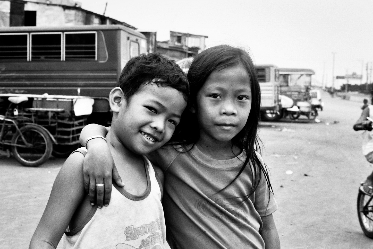 Two young children posing together outdoors with buildings and motorcycles visible in background