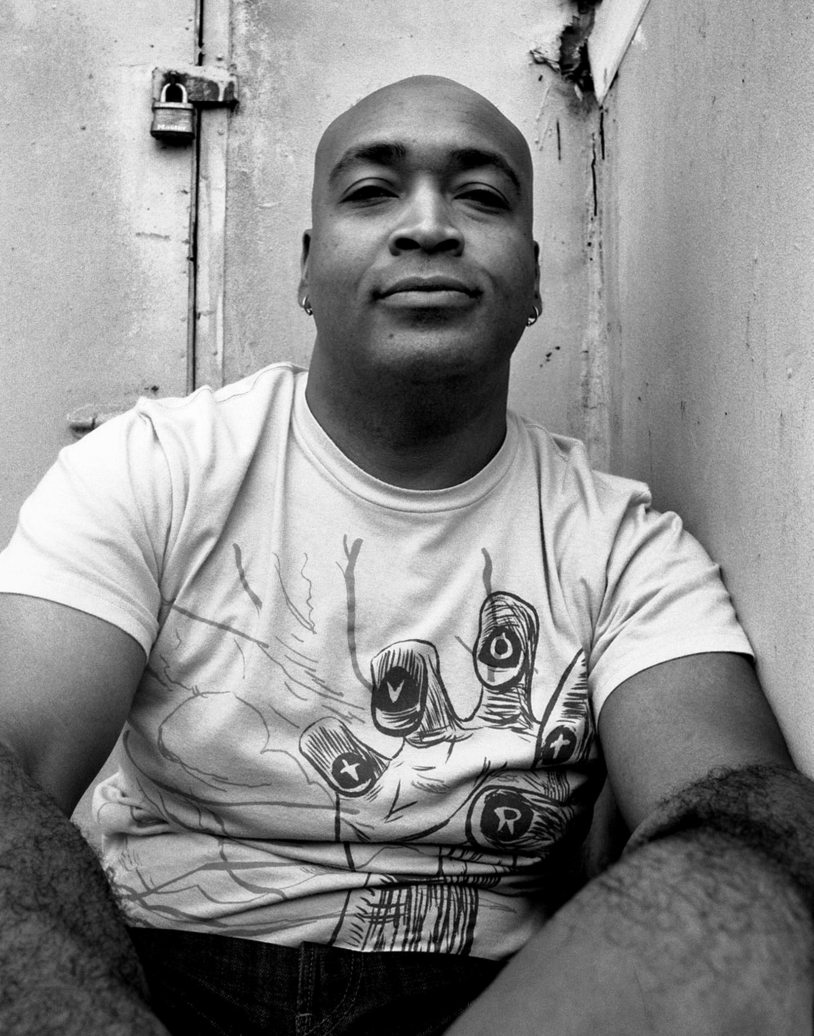 Portrait of bald man in graphic t-shirt with hand illustration leaning against wall with padlock