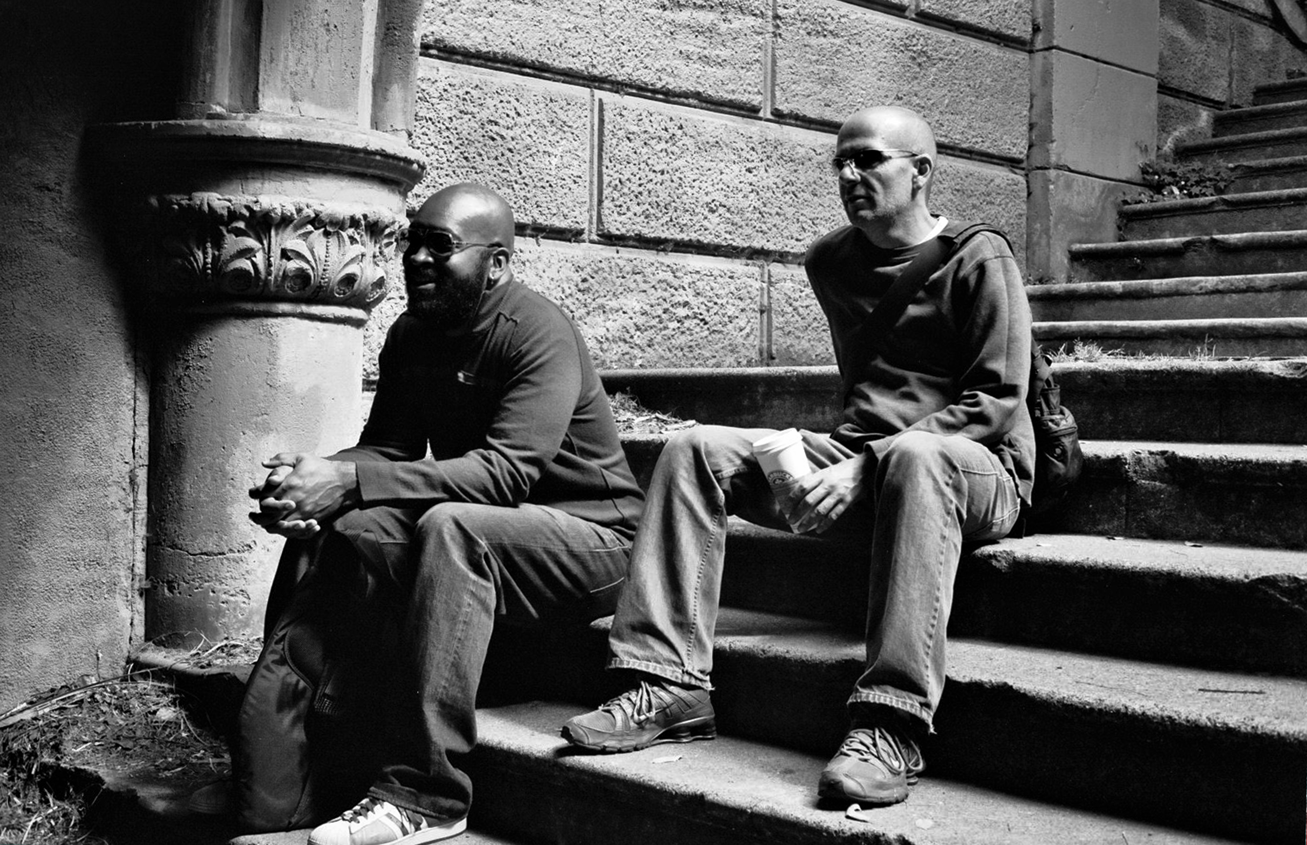 Two men sitting on stone steps next to ornate classical column in conversation wearing sunglasses
