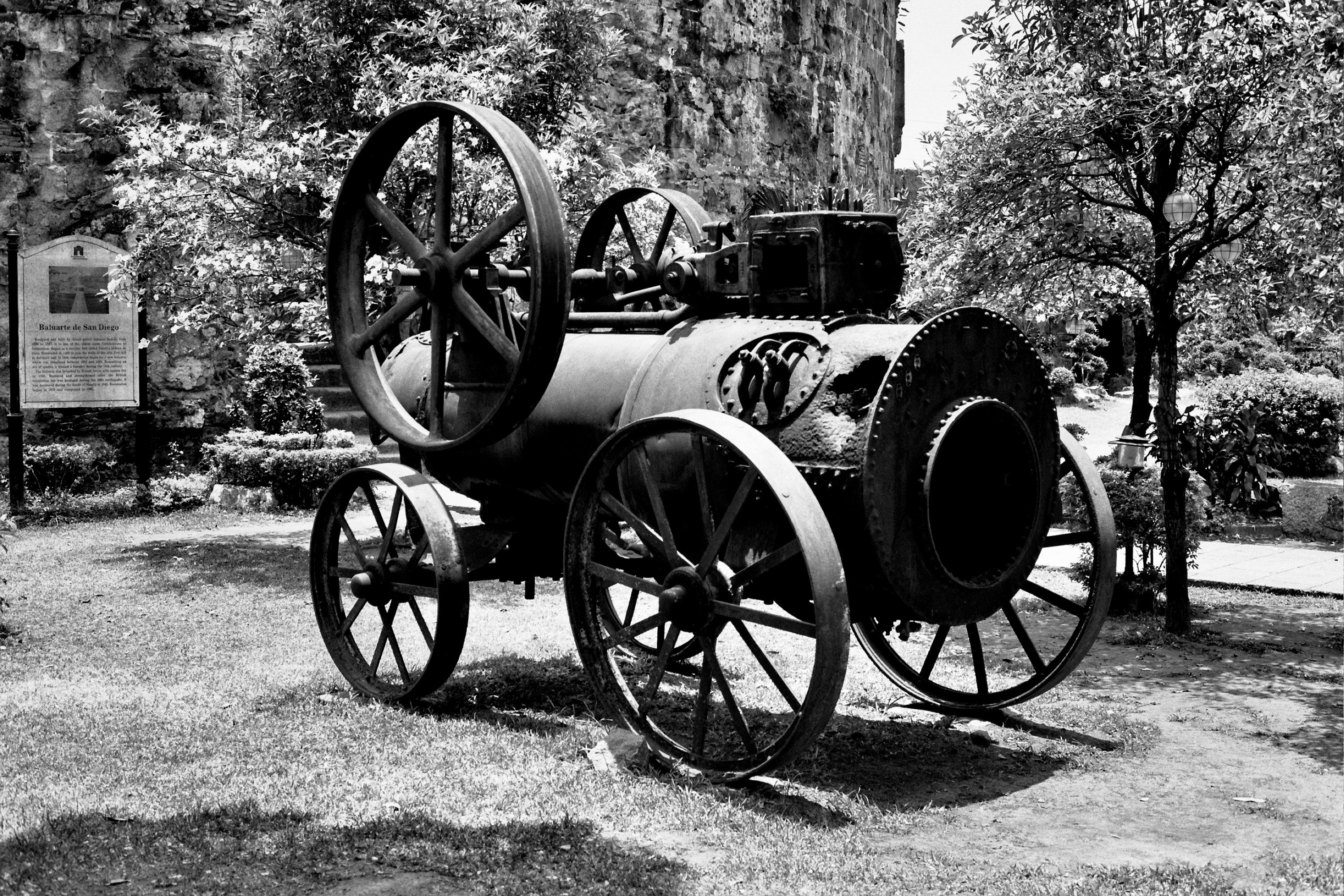 Antique steam engine machinery with large wheels and boiler displayed in historical site with trees and sign