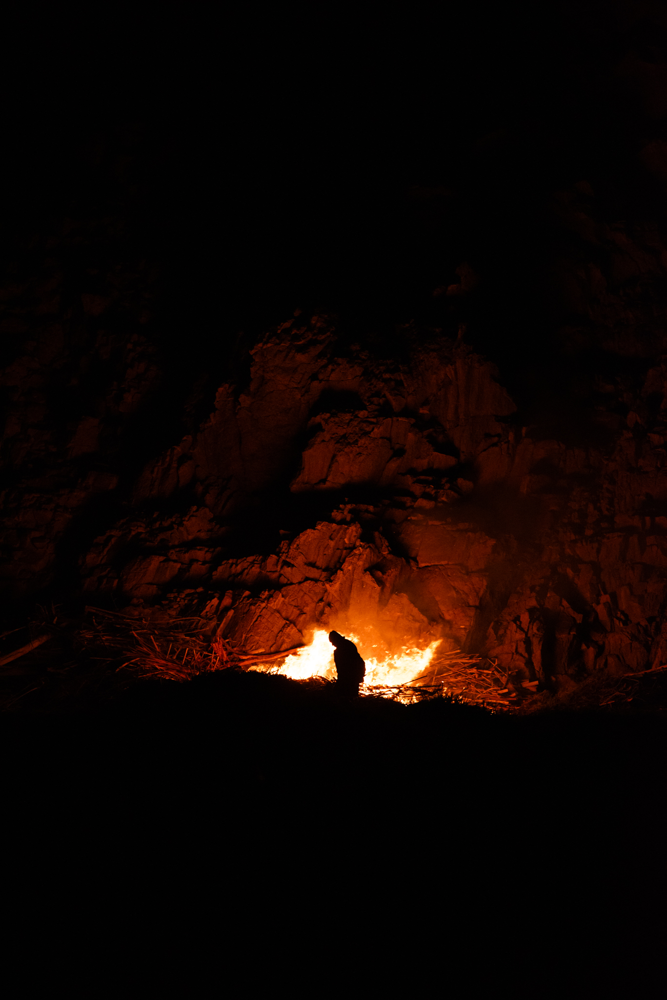 Person silhouetted by campfire at night against rocky backdrop