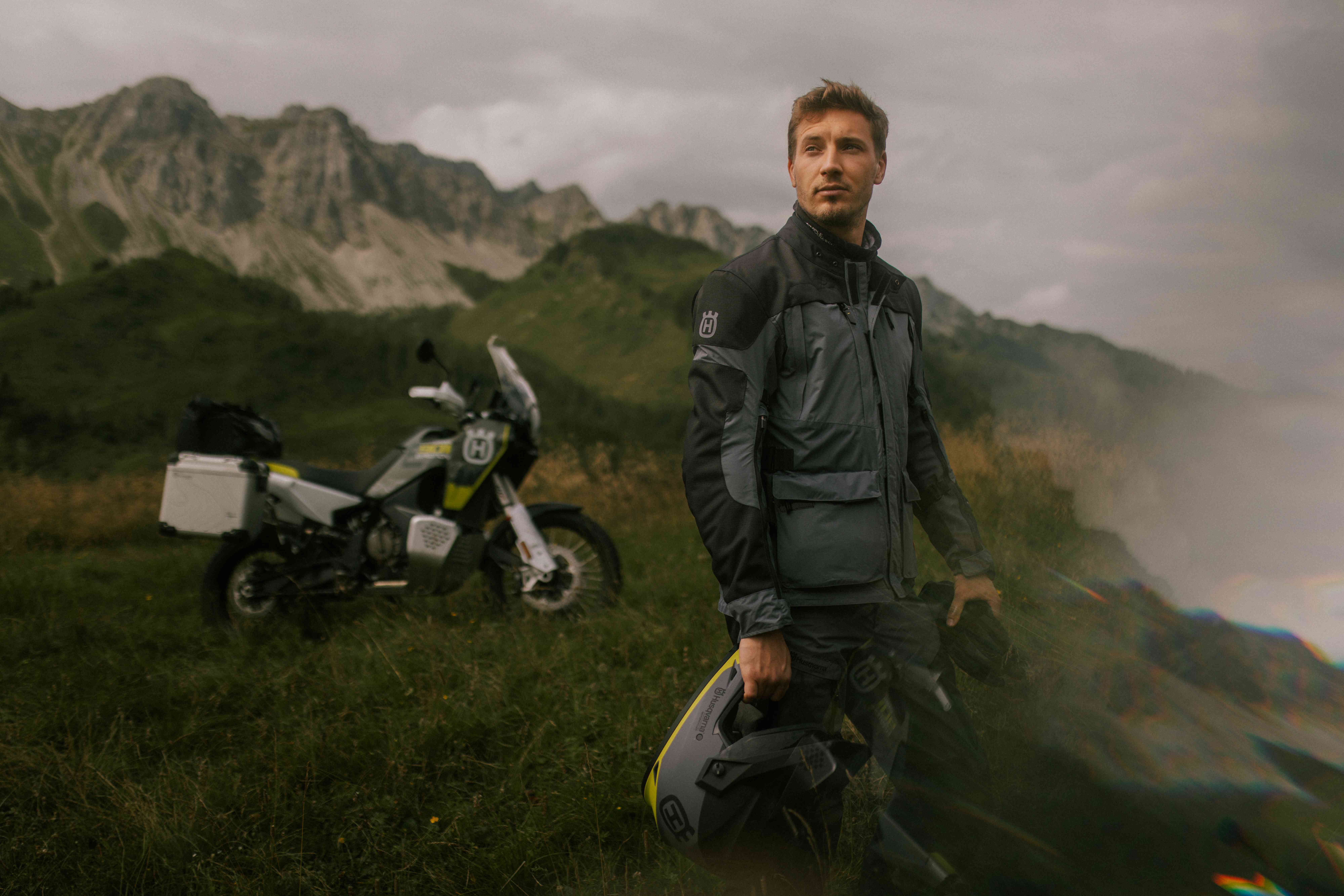 Rider in Husqvarna adventure gear holding a helmet with an alpine peak and adventure bike behind