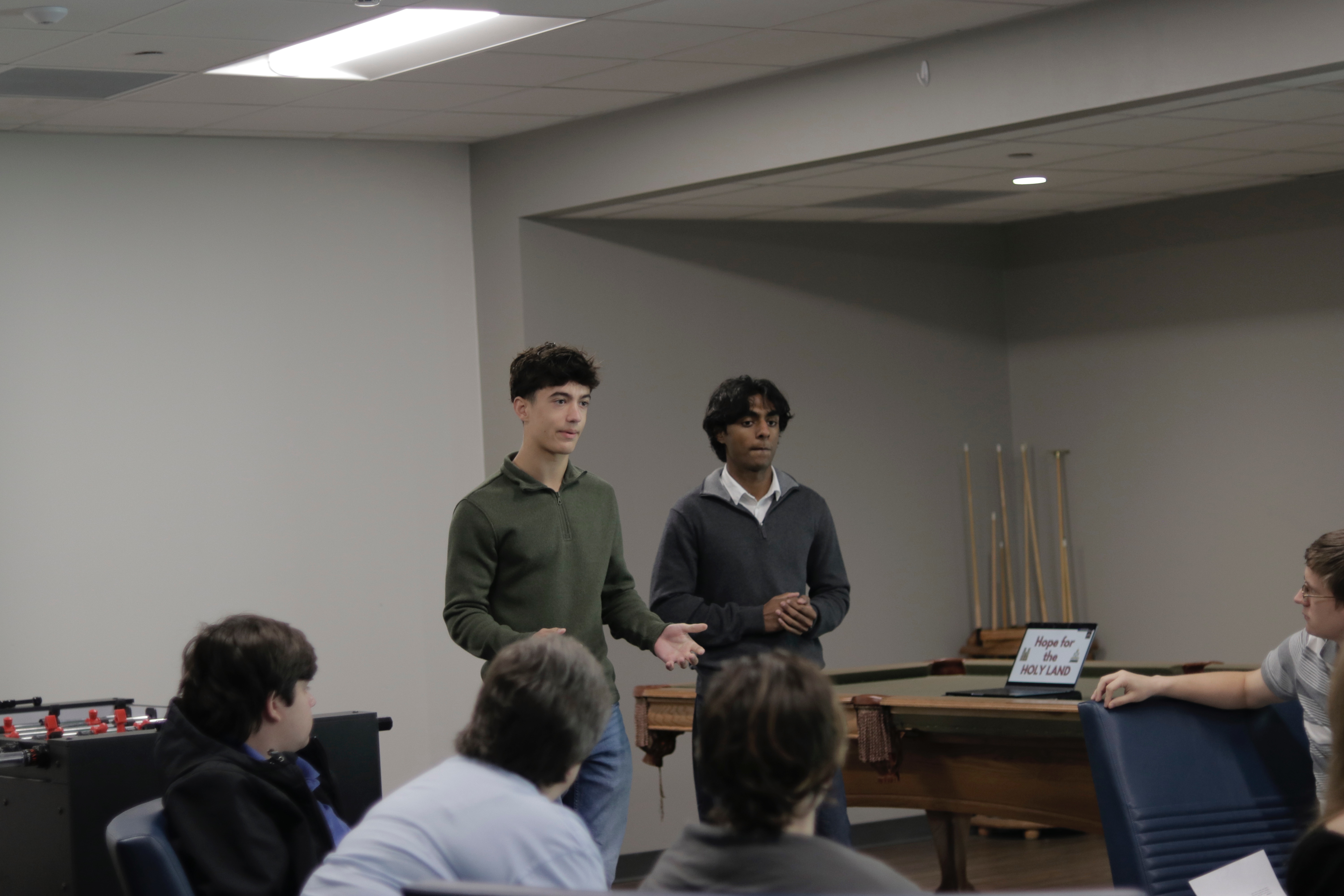 The founders speaking to a youth group beside a Hope for the Holy Land title screen
