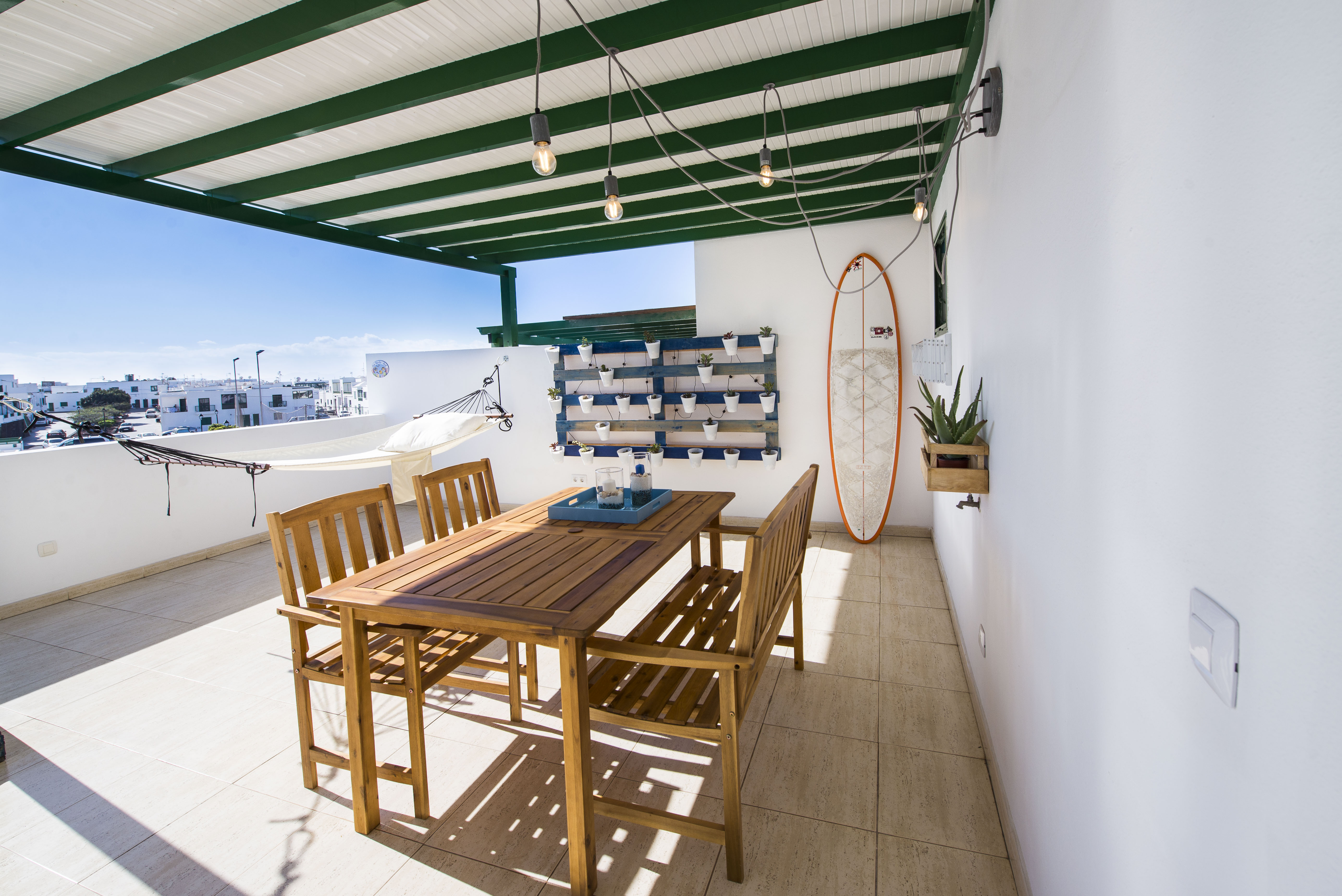 Terrace dining table under string lights with surfboard and plant wall