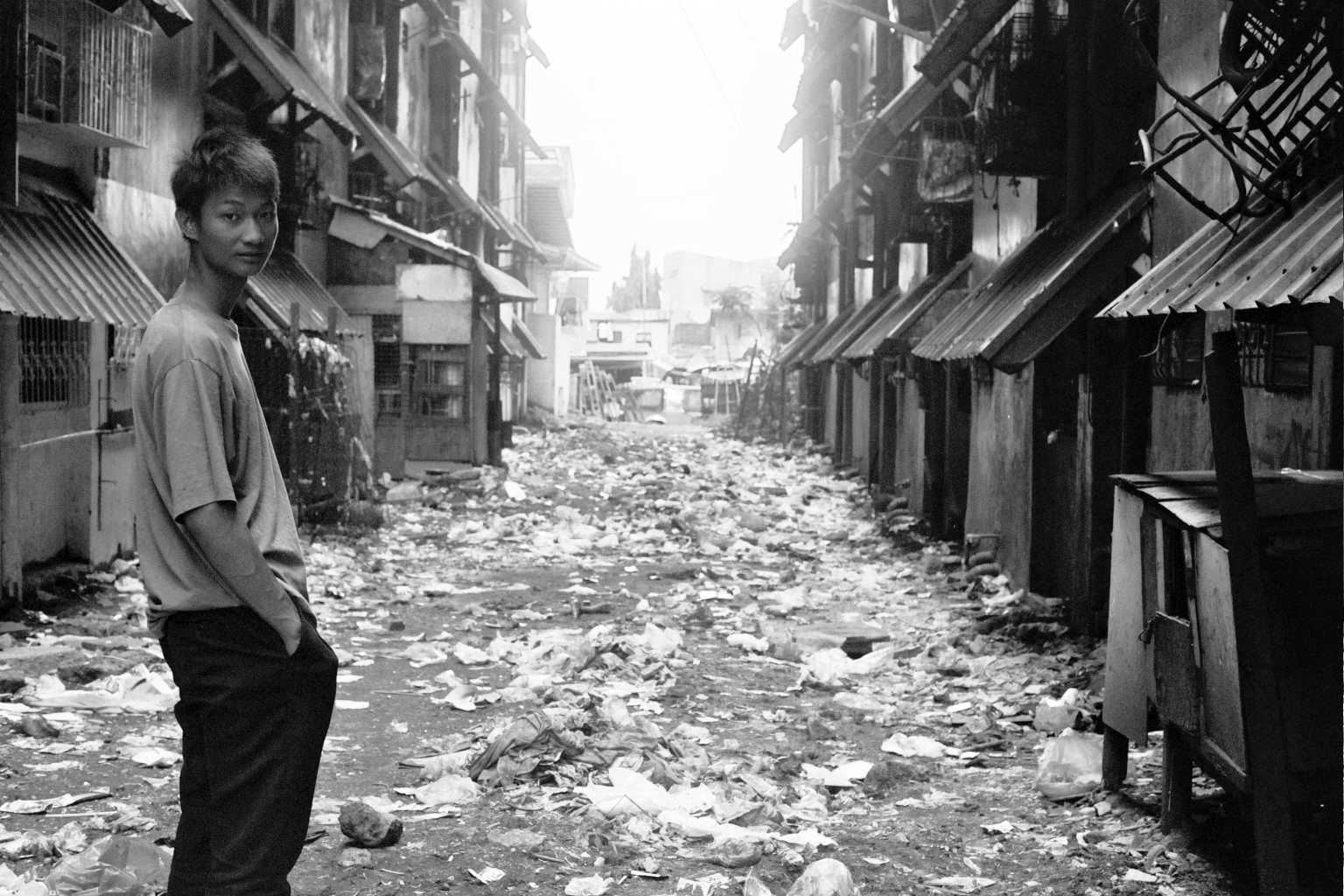 Young man standing in narrow alley with makeshift buildings and scattered garbage on ground looking back at camera