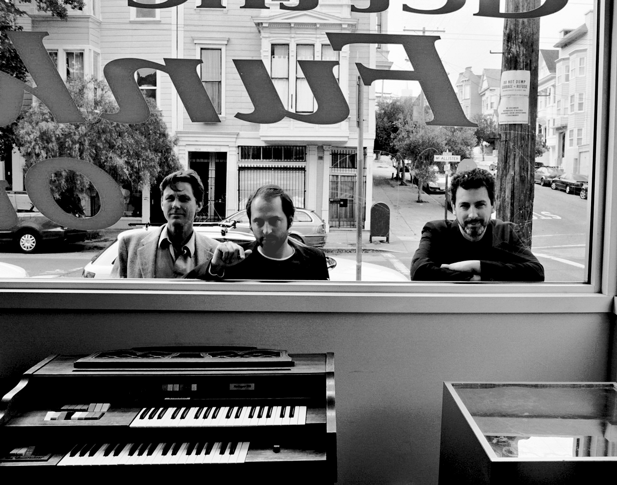 Three men posing through storefront window with illuminated large numbers on building exterior keyboards visible in foreground
